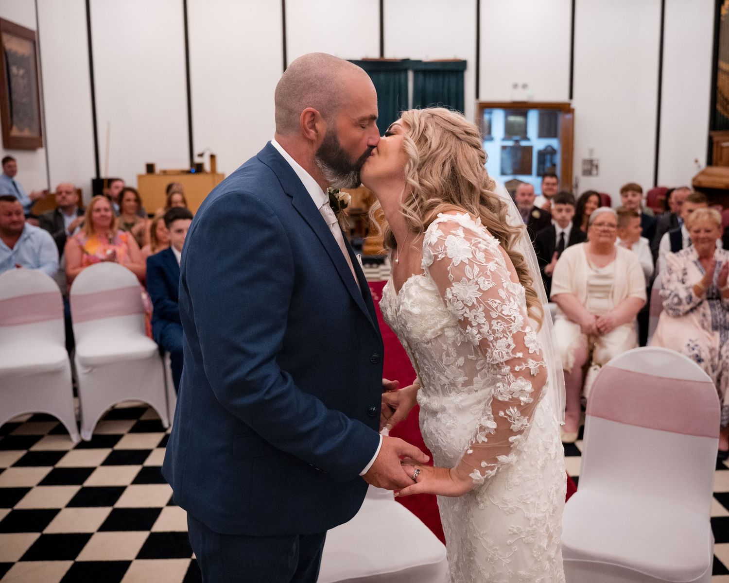 Bride and groom share first kiss at wedding ceremony in elegant hall with guests seated in white chairs.