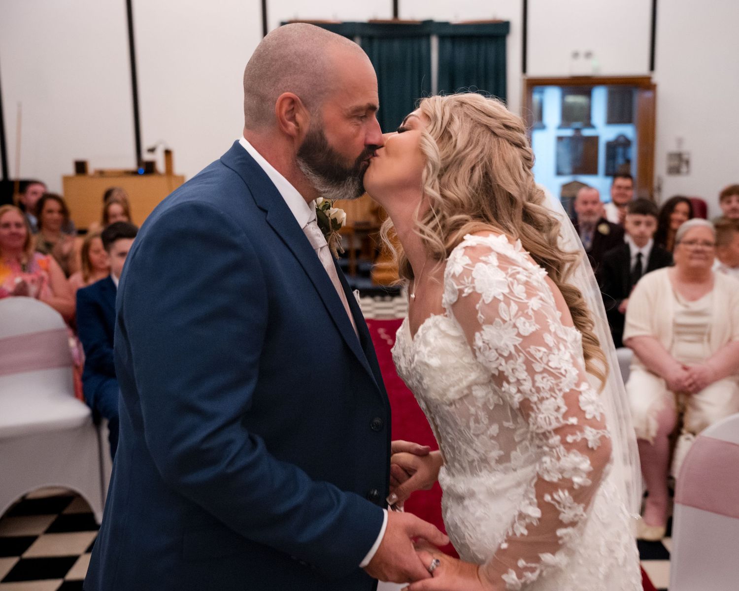 Bride and groom share first kiss at wedding ceremony with guests seated in background.