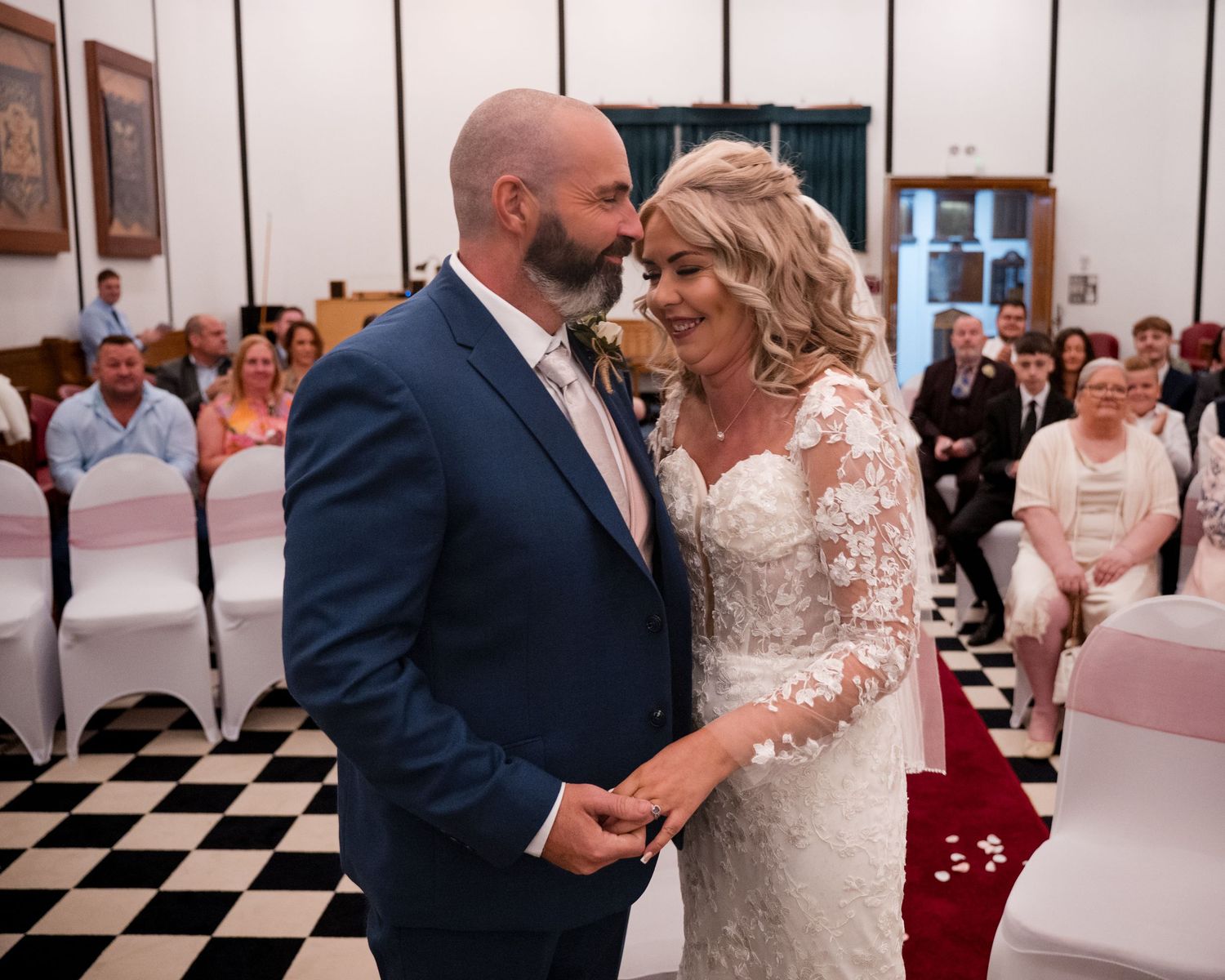 Bride and groom share a tender moment at their wedding ceremony in an elegant venue with guests seated in white chairs.