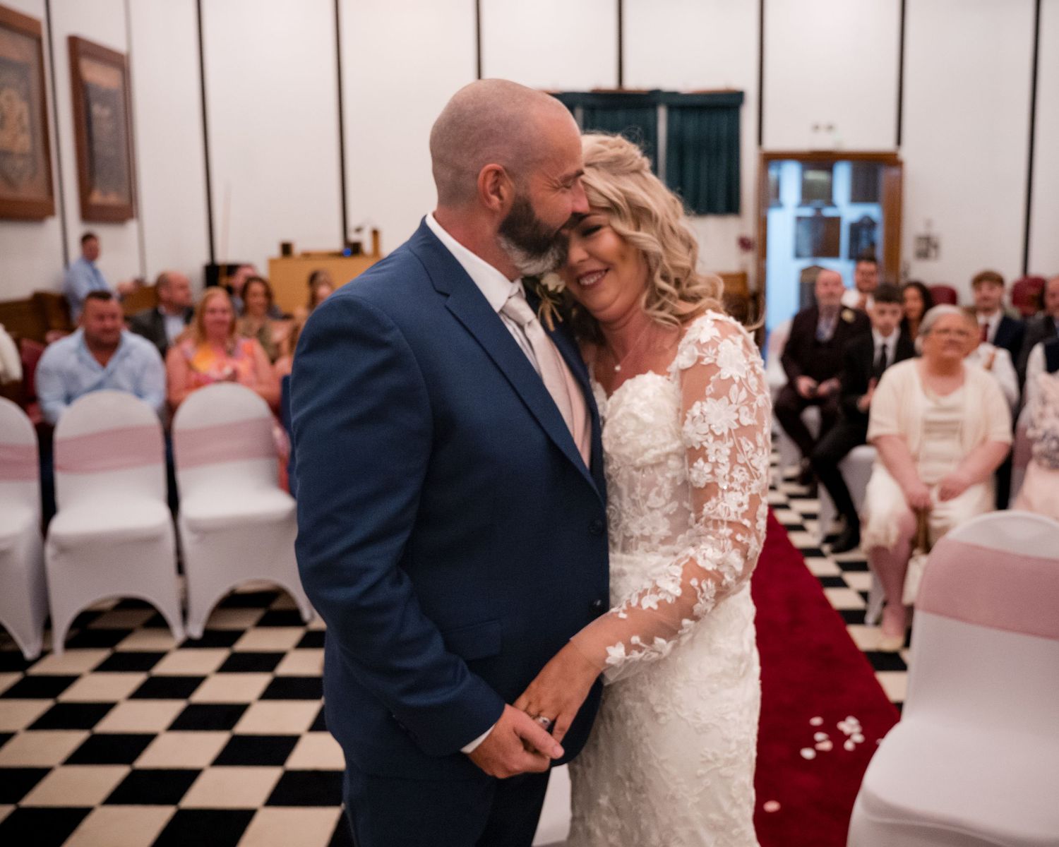 A bride and groom share a tender first kiss at their wedding ceremony in a elegant venue with guests seated behind them.