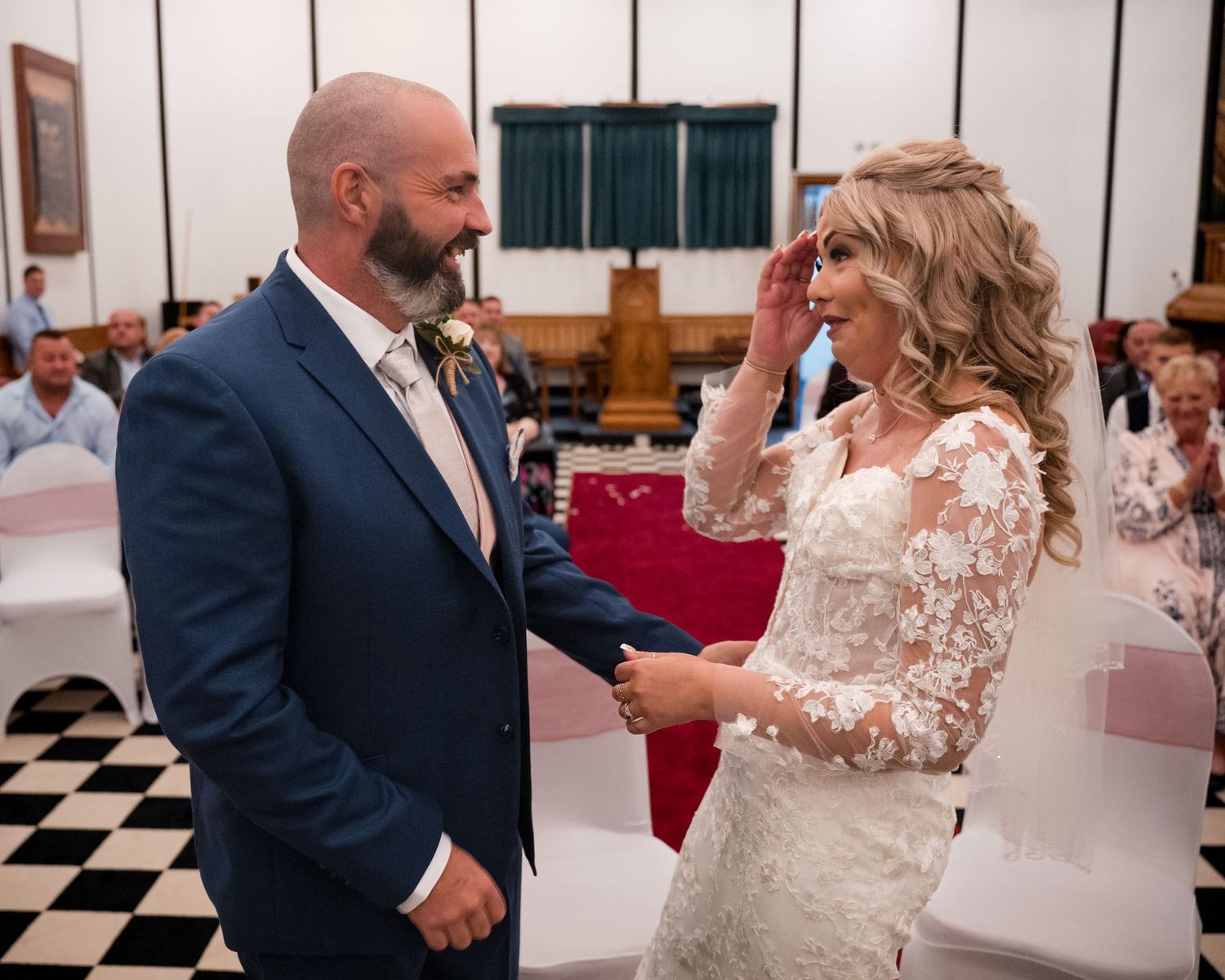 Bride in lace gown and groom in blue suit hold hands during emotional wedding ceremony in decorated hall.