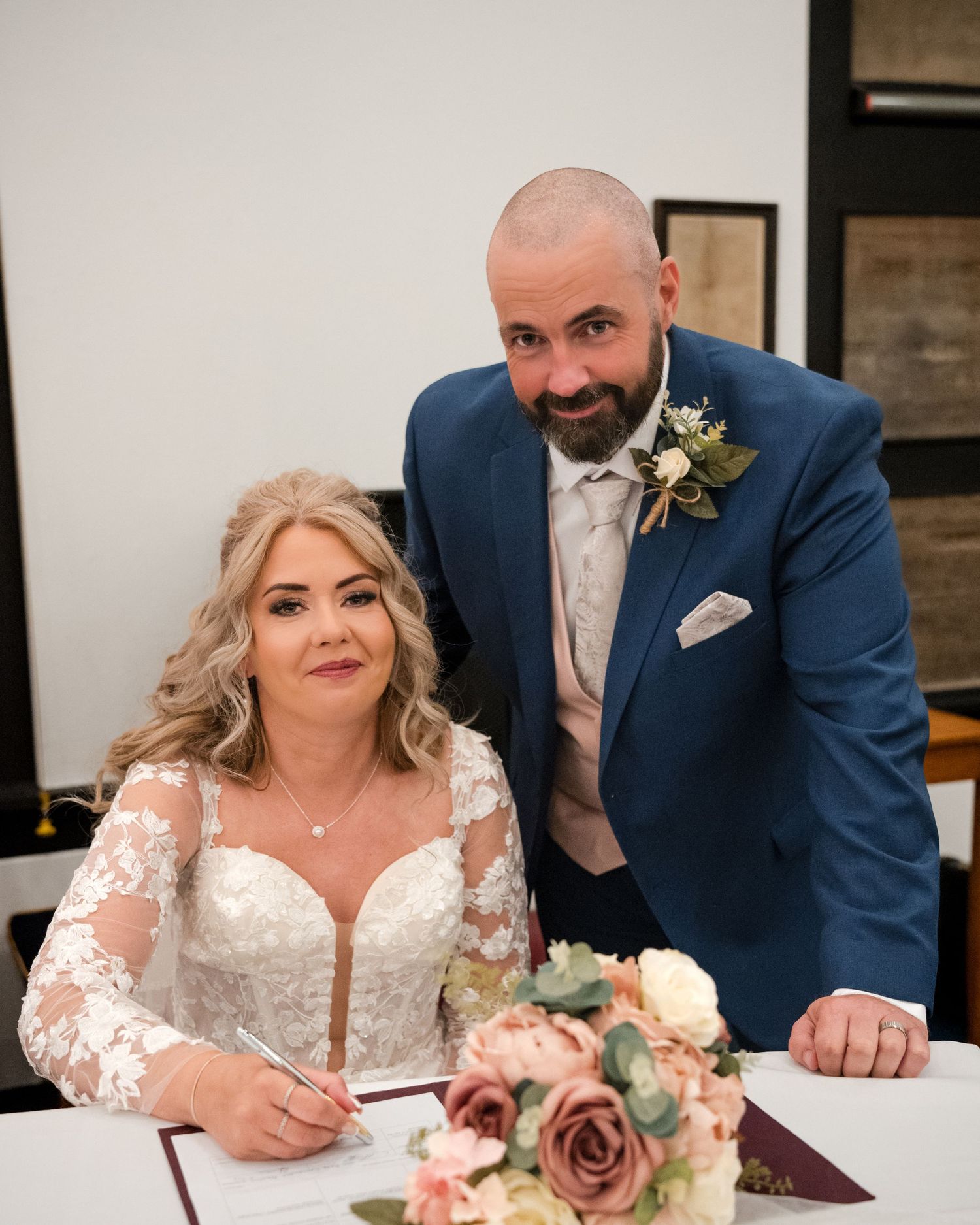 Bride and groom signing wedding register, bride in lace gown, groom in blue suit with floral boutonniere.