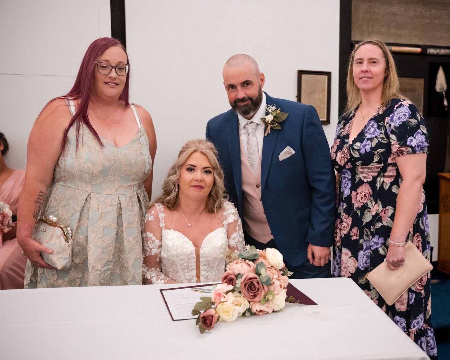 Bride seated at signing table with bouquet, groom in blue suit, and two women witnesses at wedding ceremony.