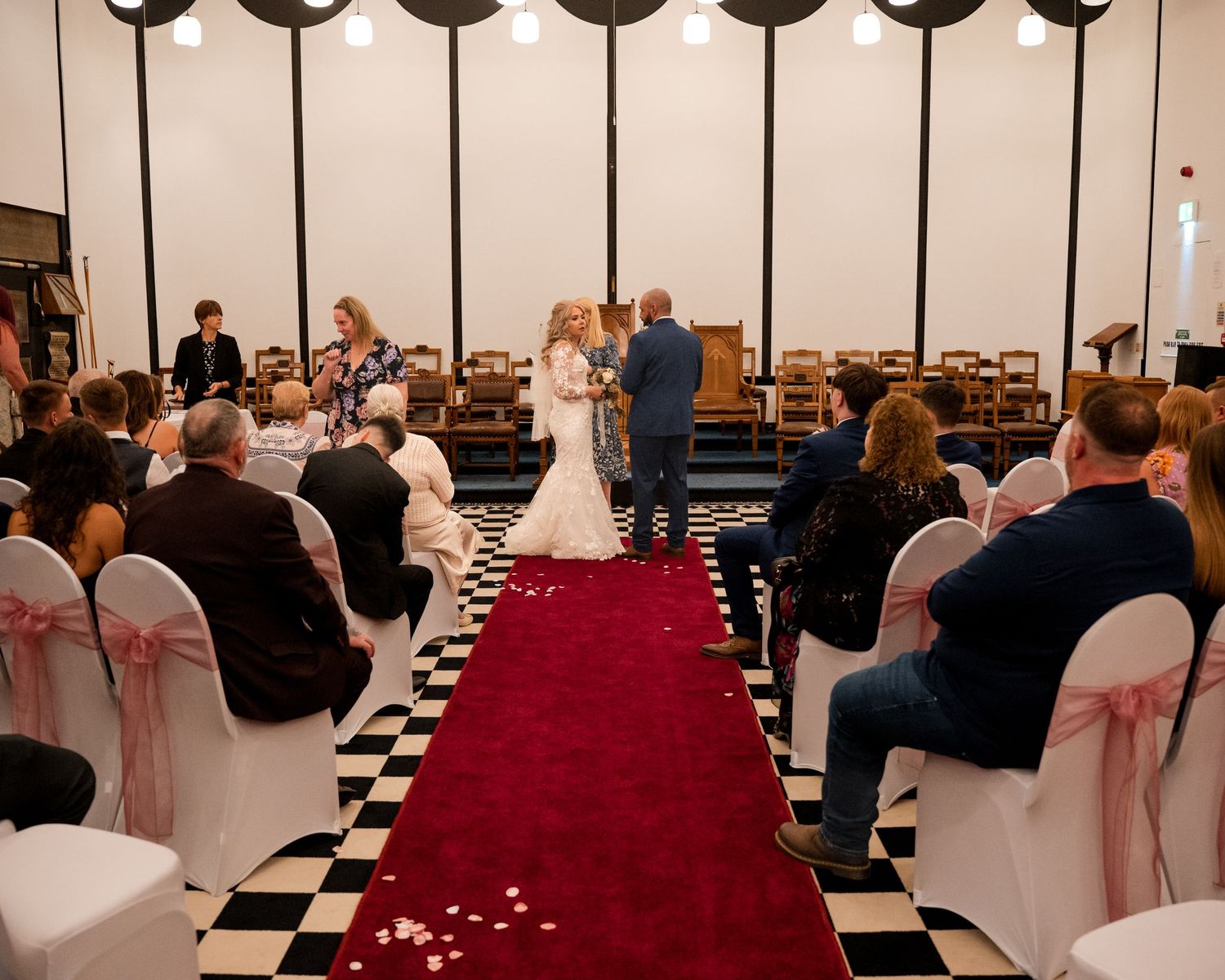 Wedding ceremony in a hall with red carpet aisle, checkered floor, white chairs with pink sashes, and guests watching the couple.