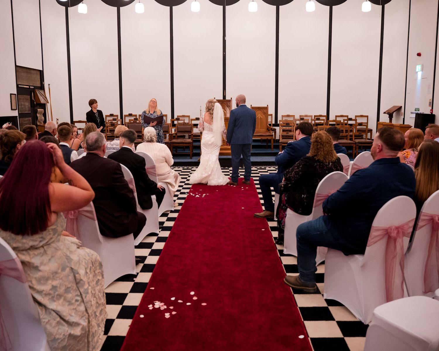 Wedding ceremony in a chapel with bride and groom at altar, red carpet aisle, checkered floor, and seated guests on white chairs.