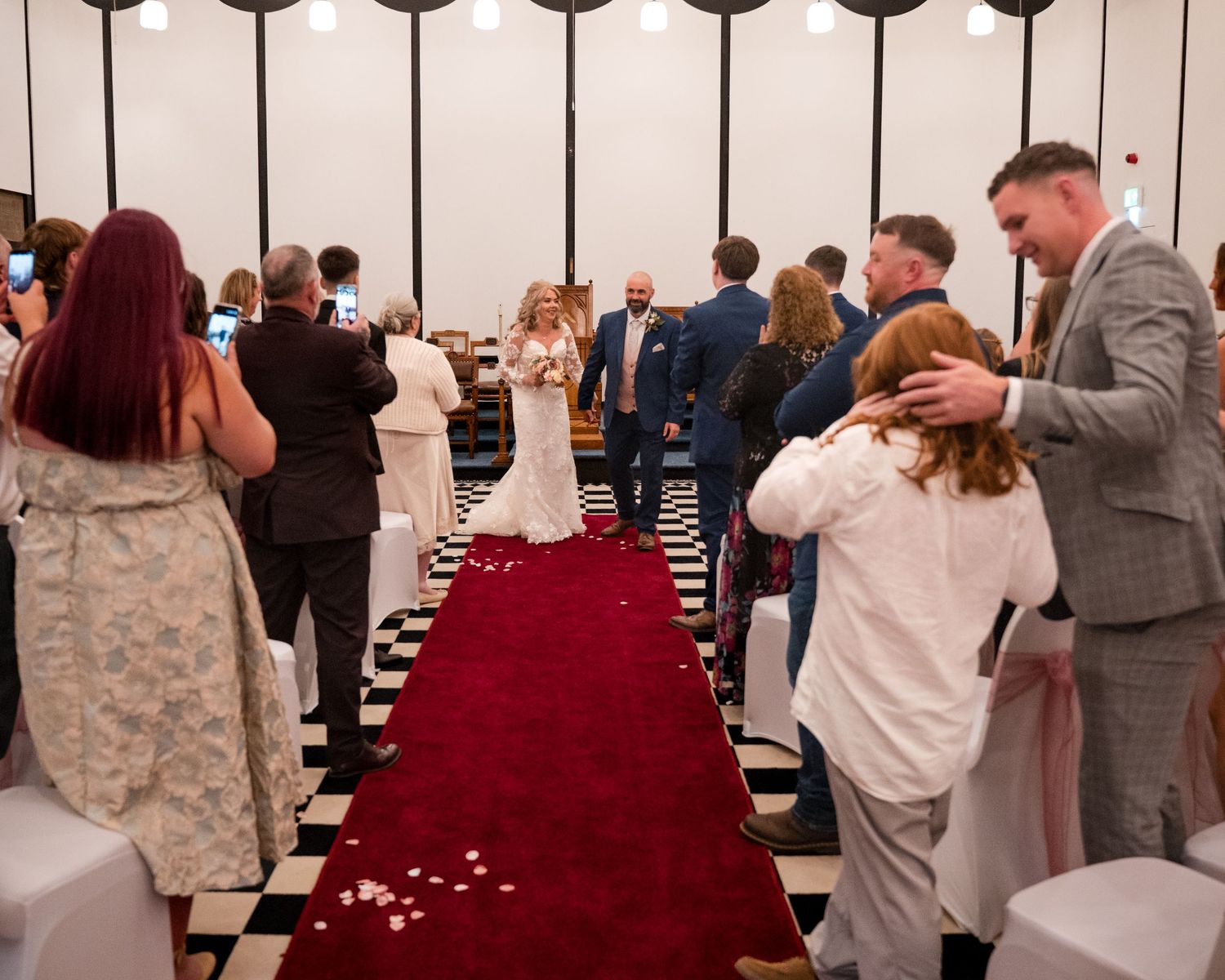 Bride walks down red carpet aisle at wedding ceremony with guests standing and cheering in elegant venue.