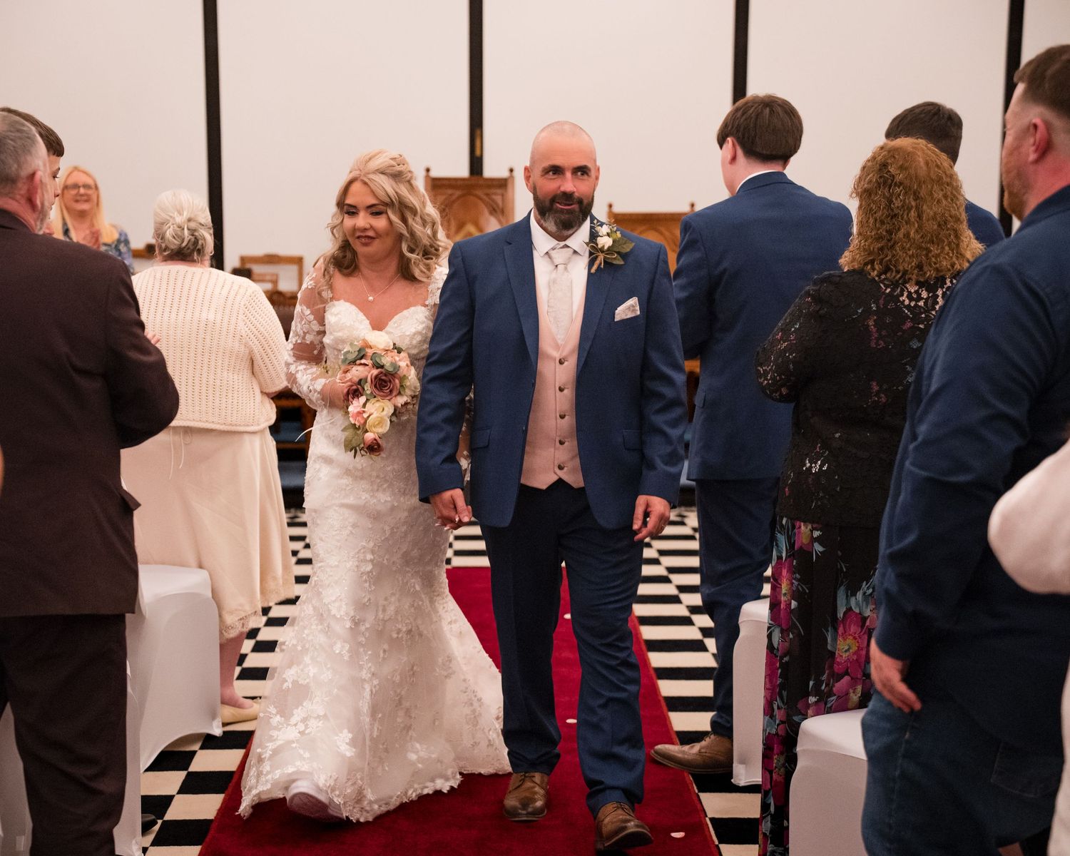 Bride and groom walking down the aisle on a red carpet in a wedding venue with checkered floors.