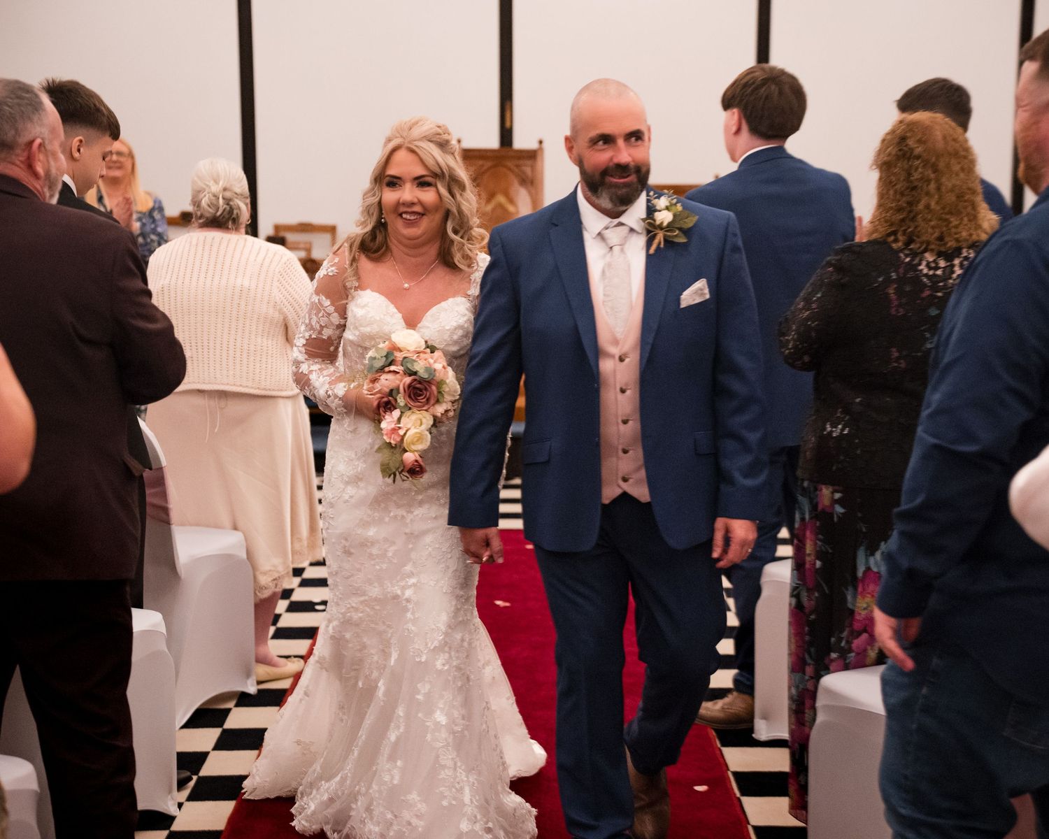 Bride in lace gown and groom in blue suit walk down the aisle smiling at their wedding ceremony.