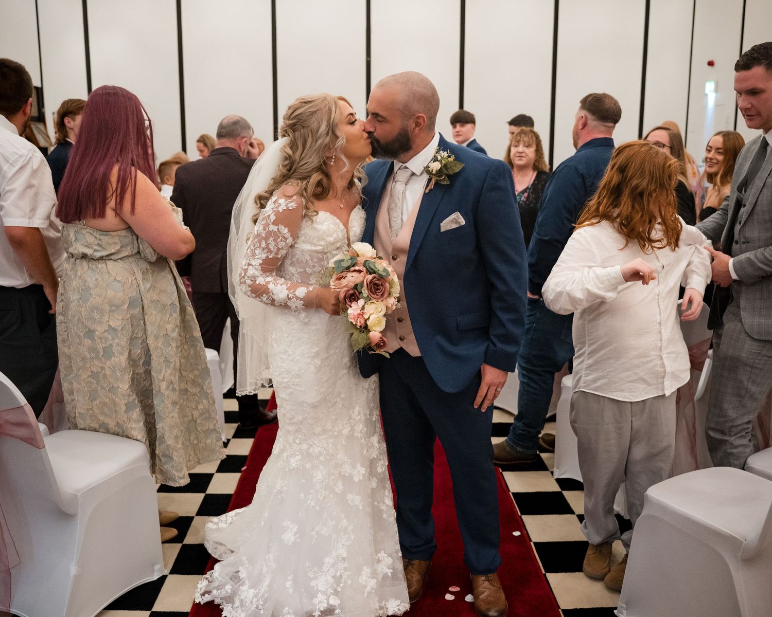 Bride and groom share first kiss at wedding ceremony on checkered floor surrounded by guests.