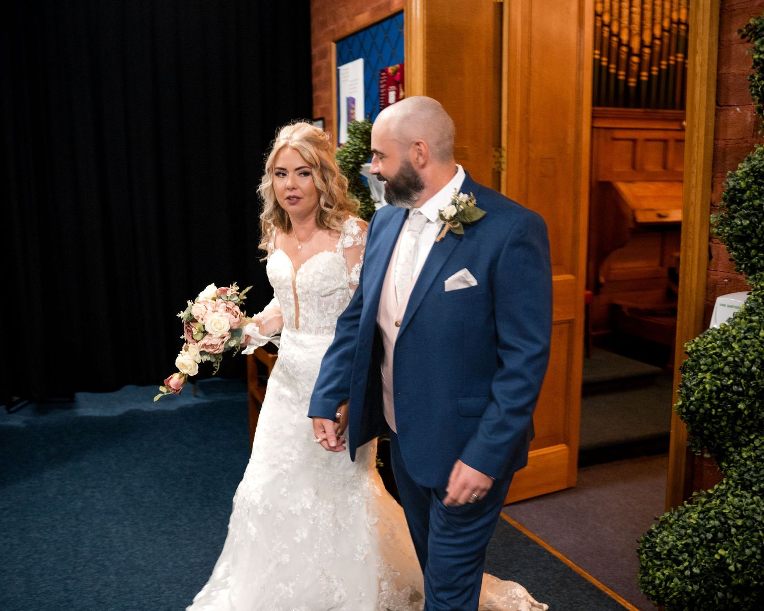 Bride in lace wedding gown holding bouquet walks with groom in blue suit after church wedding ceremony.