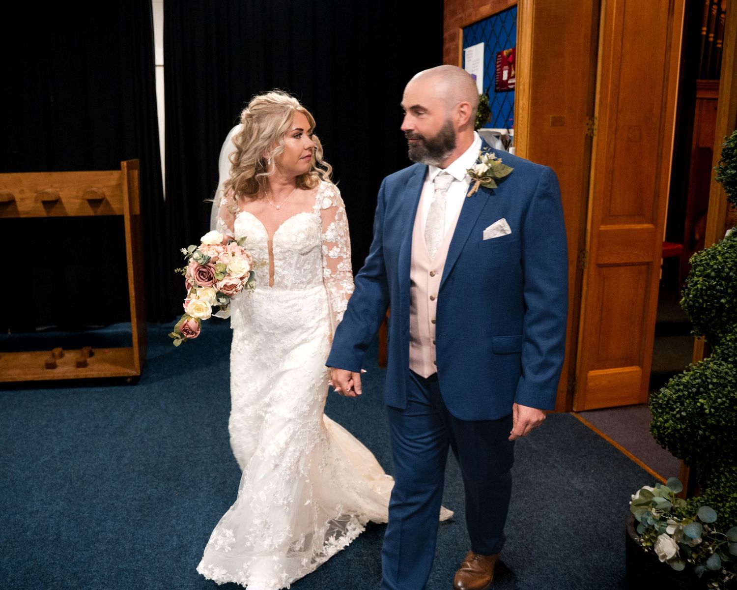 Bride in lace gown holding bouquet walks hand-in-hand with groom in blue suit during wedding ceremony.