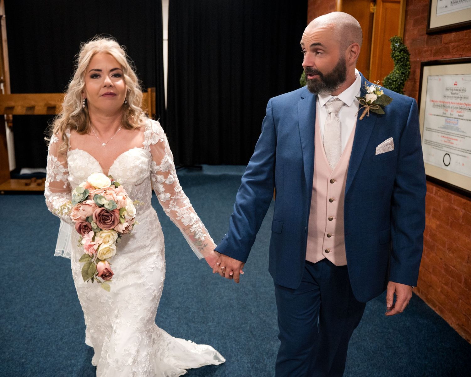 Bride in lace gown and groom in blue suit hold hands after wedding ceremony.