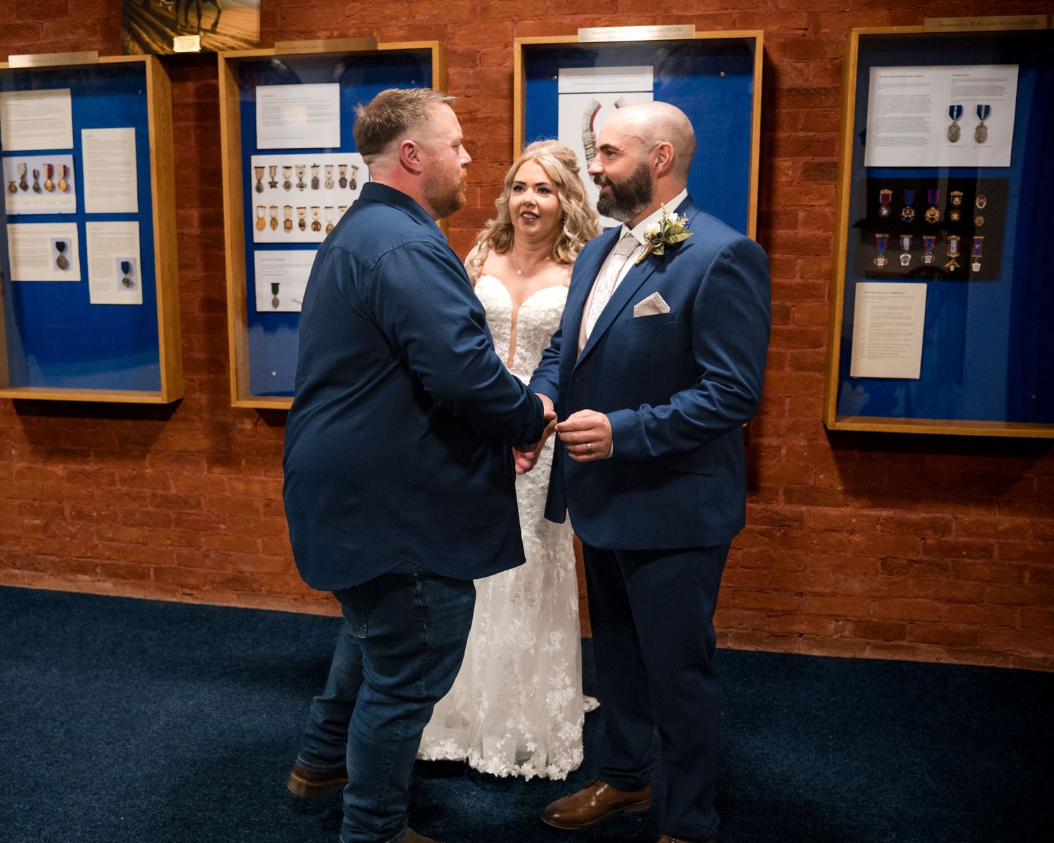 Bride and groom share a joyful moment with a guest in a navy suit inside a wedding venue with display cases.