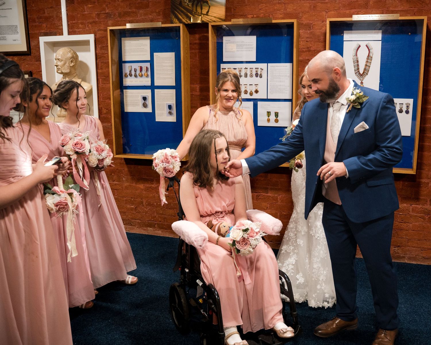 Groom in blue suit with bride in wheelchair surrounded by bridesmaids in pink dresses holding bouquets at wedding.