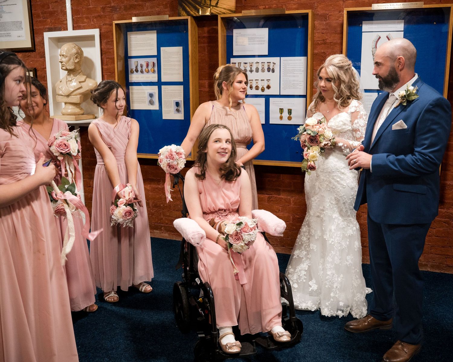 Wedding party with bride in white gown, groom in navy suit, and bridesmaids in pink dresses, one bridesmaid seated in wheelchair.