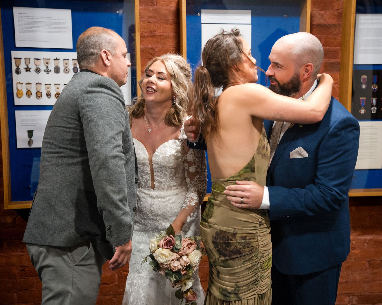 Bride in white lace gown holds bouquet while guests embrace at a wedding reception near a military medals display.