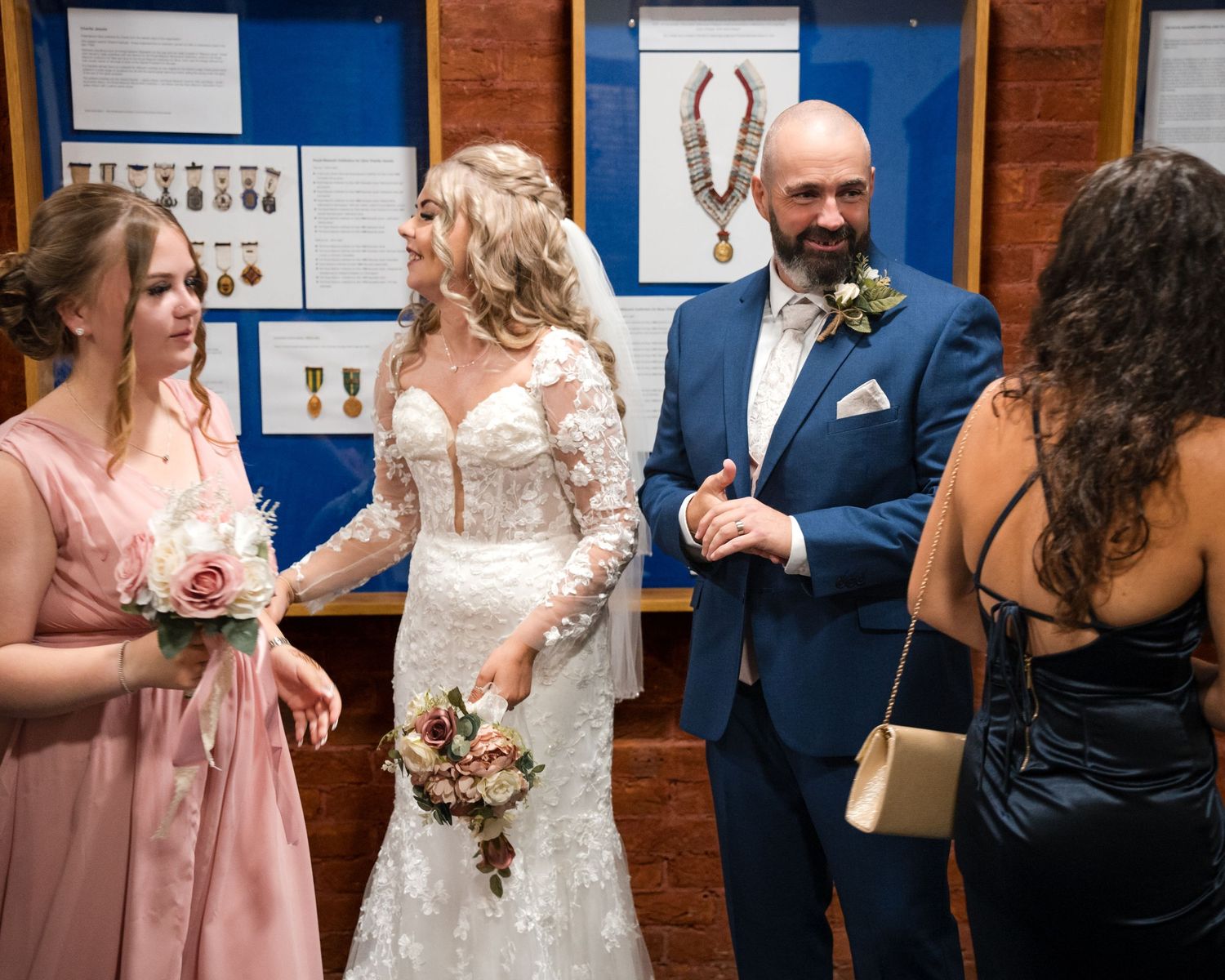 Bride in lace gown and groom in blue suit chat with bridesmaids at wedding reception near display case.
