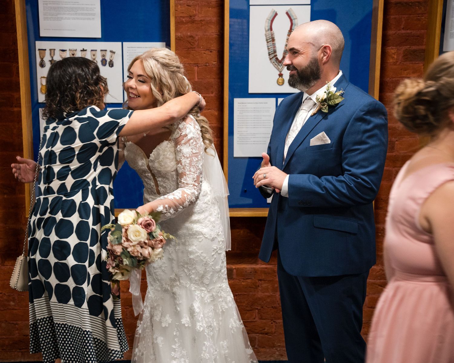 Bride and groom celebrate with guests at wedding reception, bride holding bouquet in lace gown, groom in navy suit.