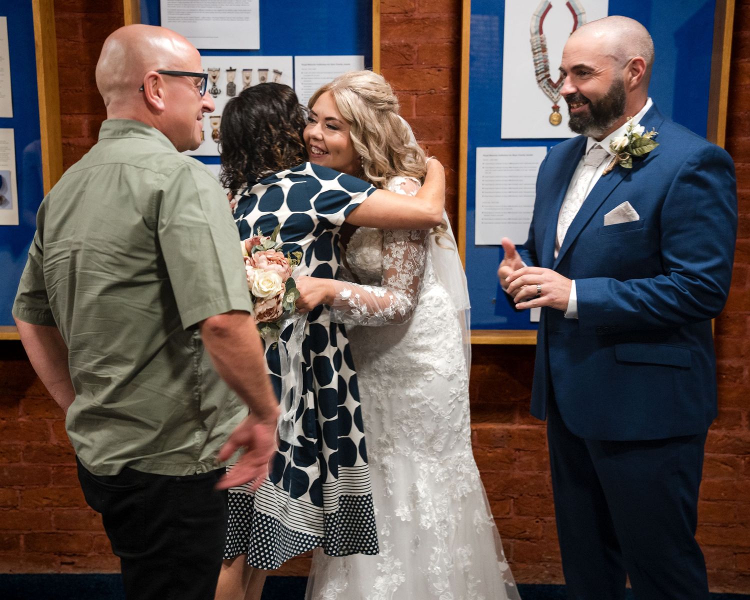 Bride in white lace gown hugging guest at wedding reception while groom in blue suit watches nearby.