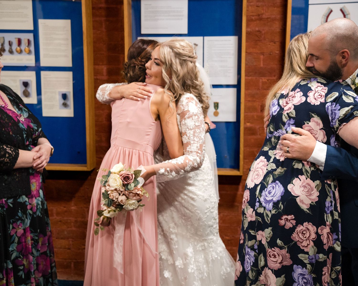 Bride in lace gown hugging bridesmaid in pink dress while guests embrace nearby at wedding reception.