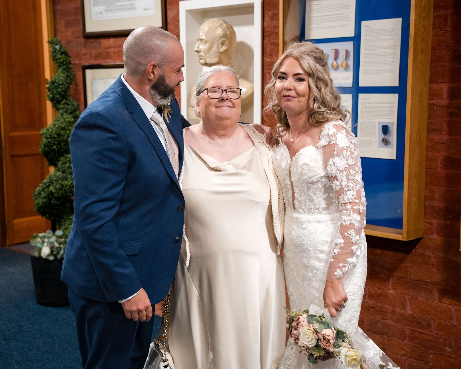 Bride and groom posing with officiant at wedding ceremony, bride wearing lace gown holding bouquet, groom in blue suit.