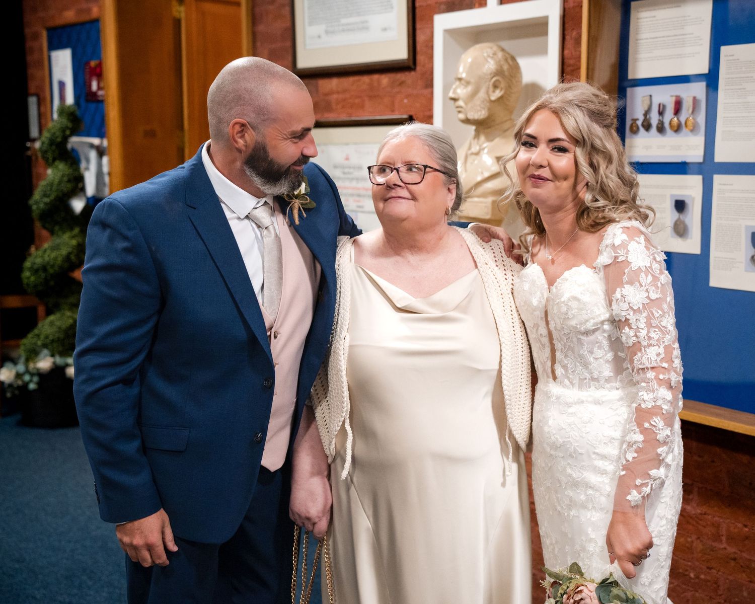 A bride in lace gown and groom in blue suit pose with an older woman in cream dress at a wedding celebration.