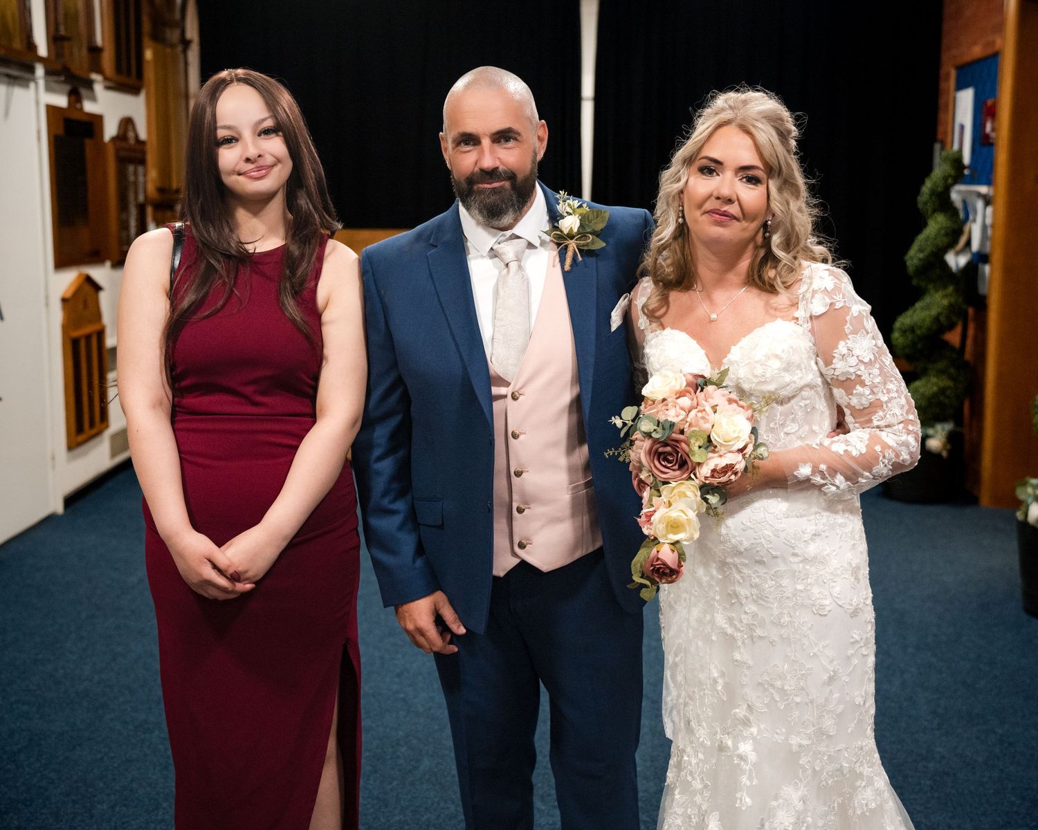 A bride in lace gown holds bouquet beside groom in blue suit and a bridesmaid in burgundy dress at a wedding ceremony.