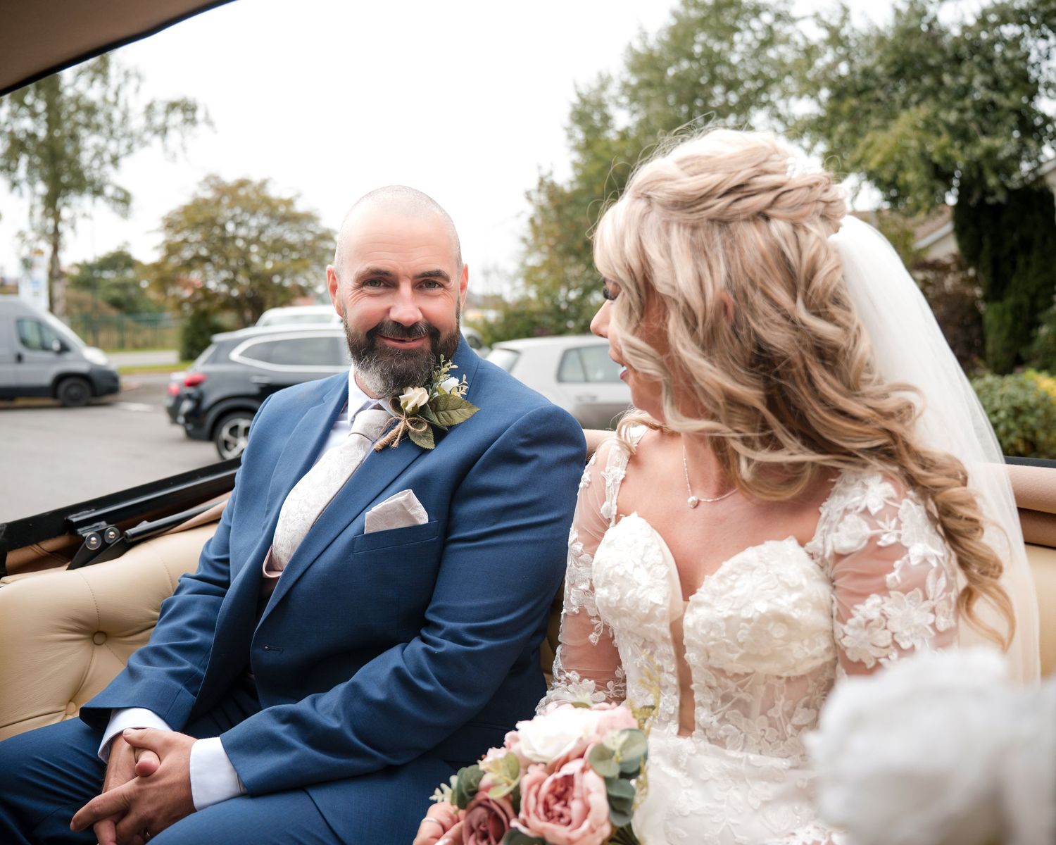Bride and groom sitting together in a vintage car, smiling, bride holding pink roses, groom in blue suit with boutonniere.