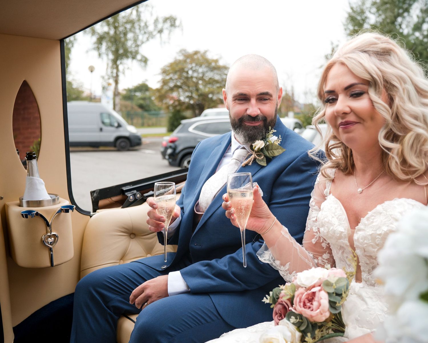 Bride and groom toasting with champagne in a vintage wedding car, groom in blue suit, bride in lace dress.