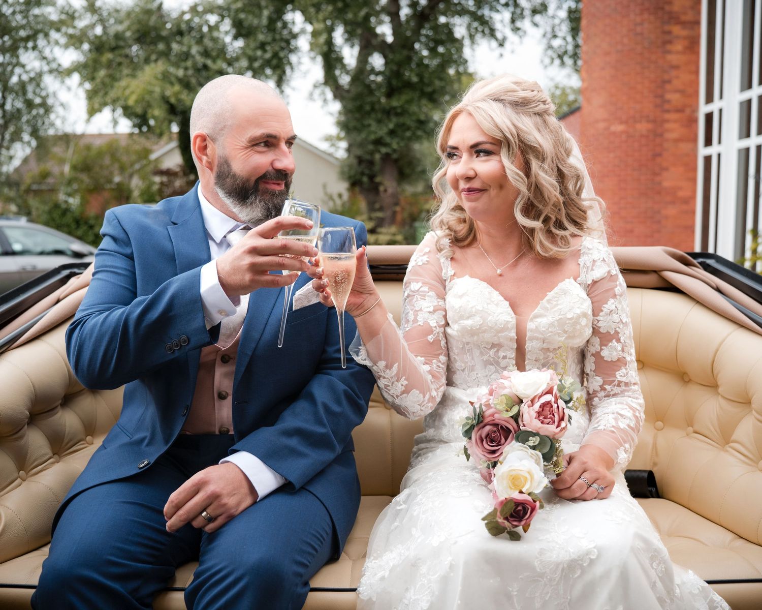 Bride and groom toasting with champagne glasses, sitting on a cream leather seat, bride holding a pink bouquet.