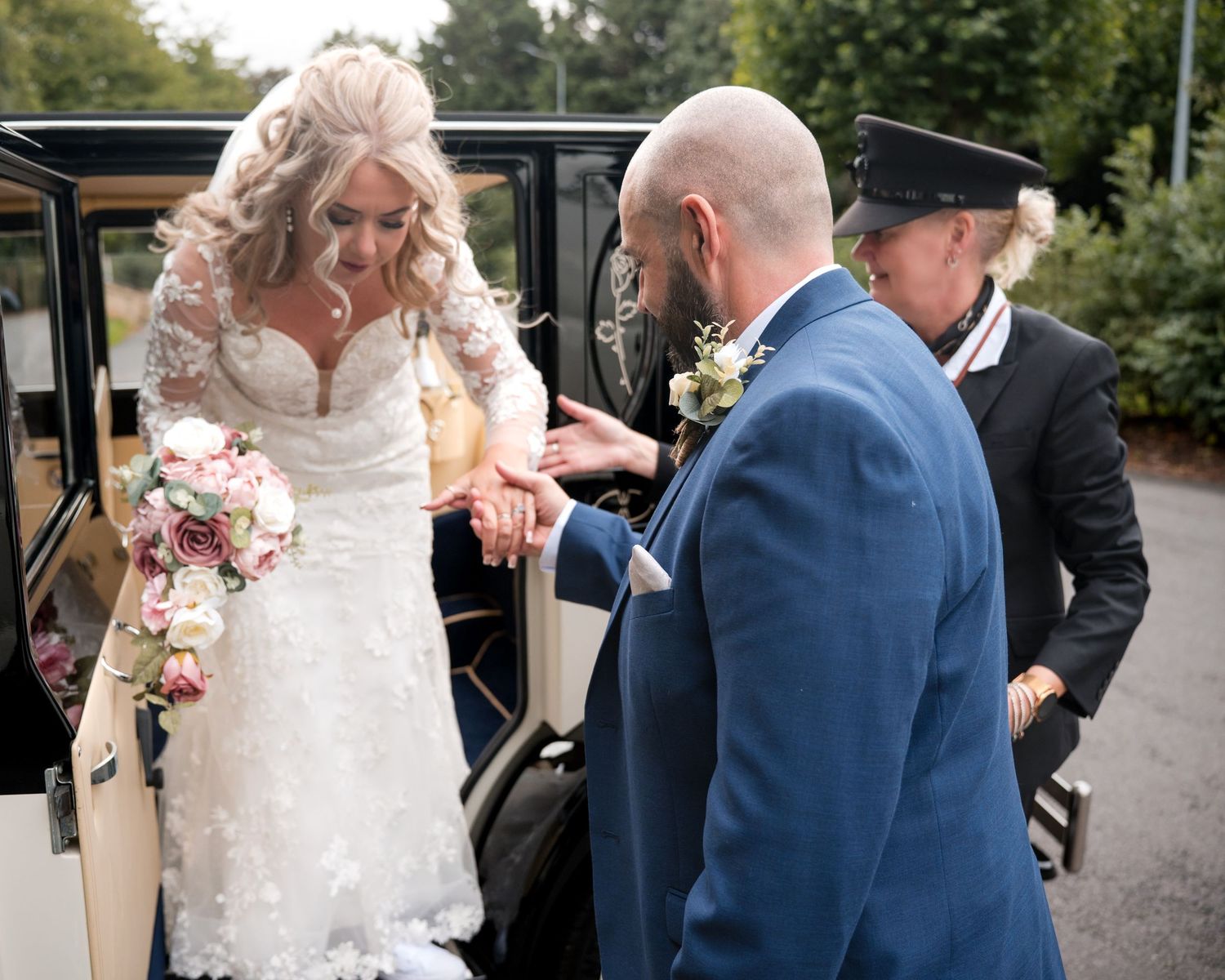 Groom in blue suit helps bride in lace wedding dress exit vintage car, holding bouquet of pink roses.