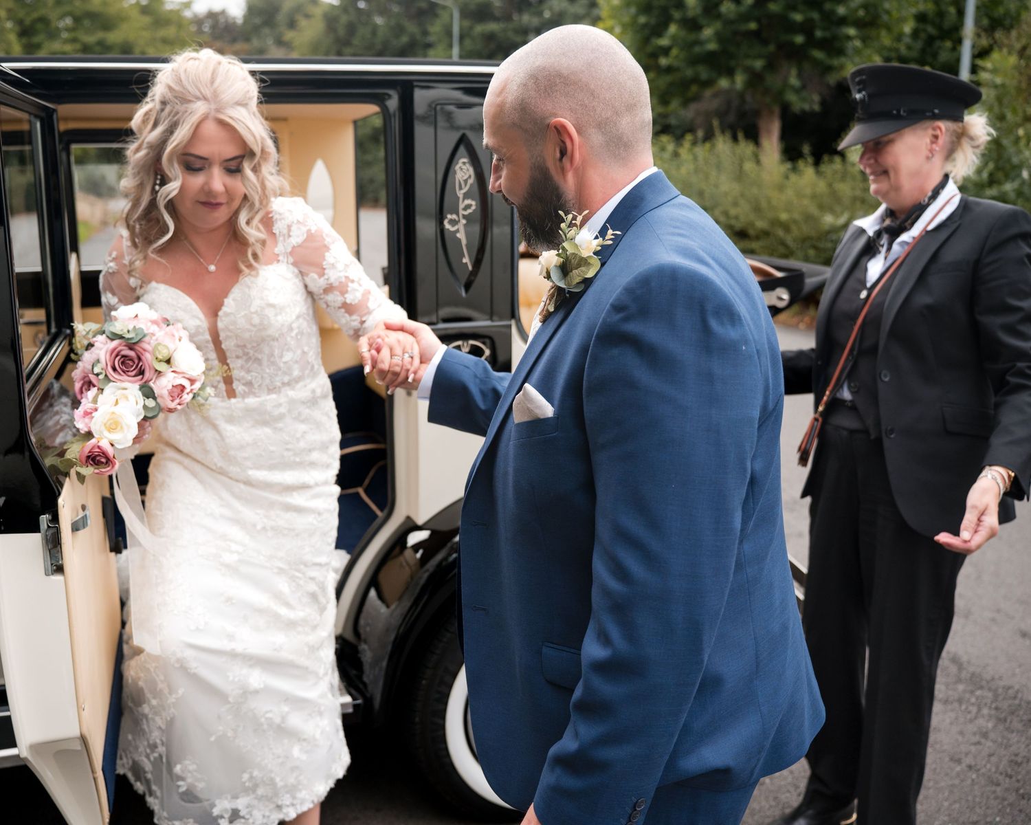 Bride in lace gown holding bouquet helped by groom in blue suit beside vintage wedding car with chauffeur.