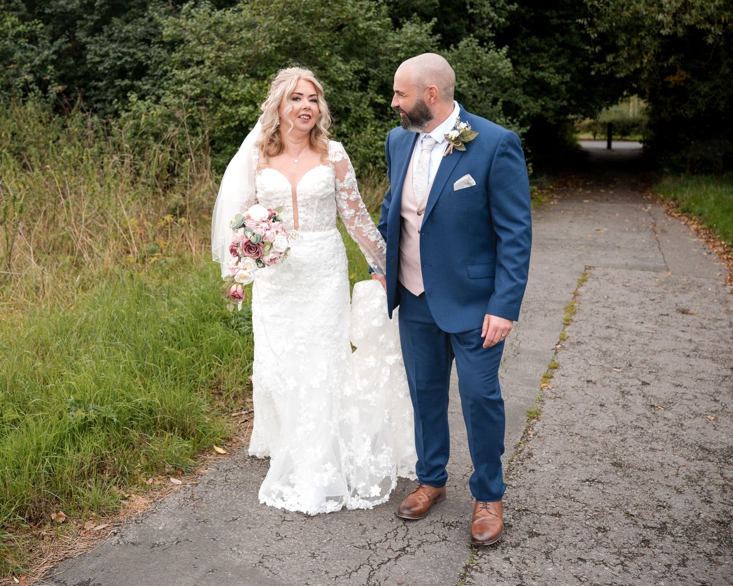 Bride in lace gown and groom in blue suit walk hand-in-hand on a tree-lined path on their wedding day.
