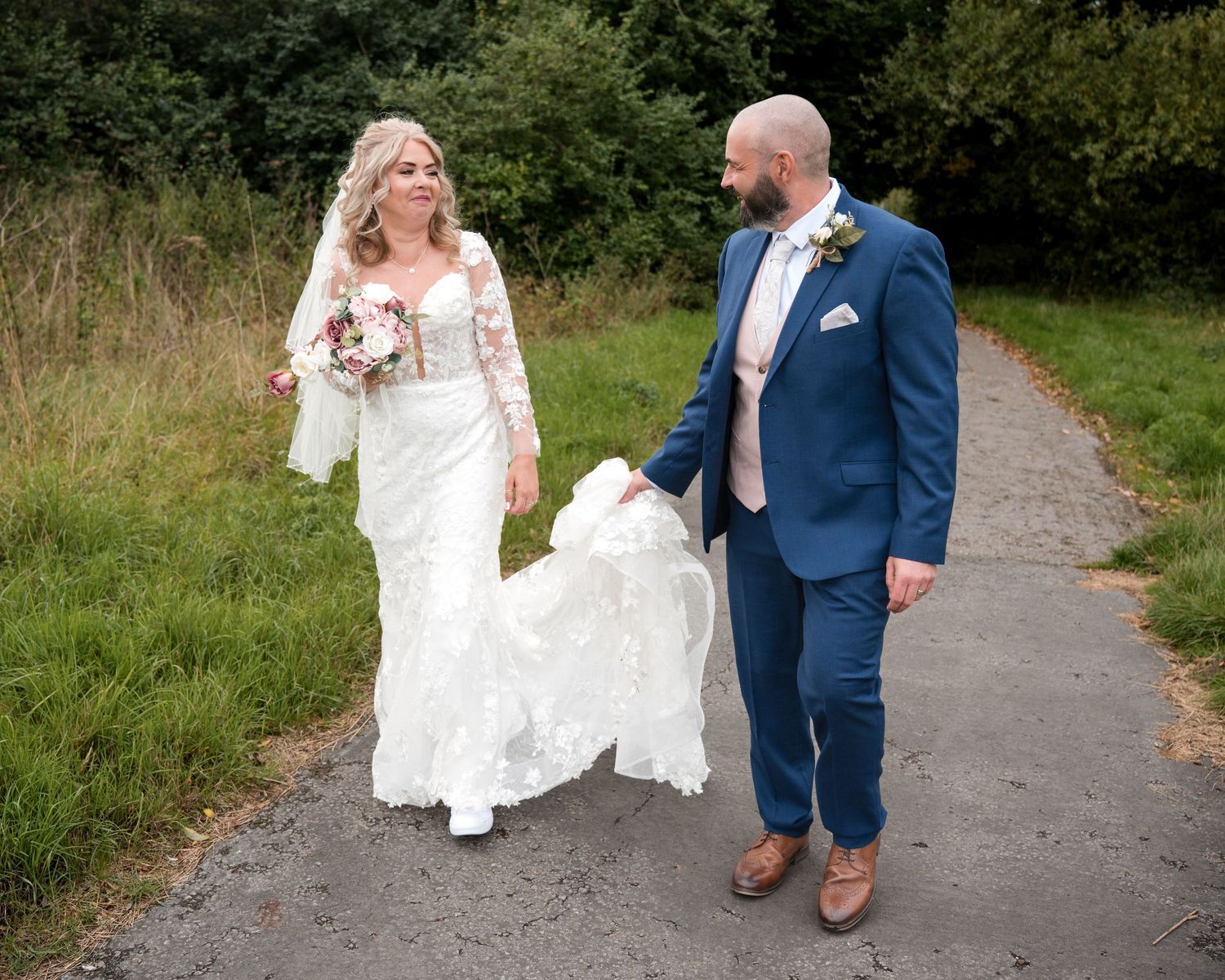 A bride in a lace gown and groom in a blue suit walk hand in hand along a path surrounded by greenery.