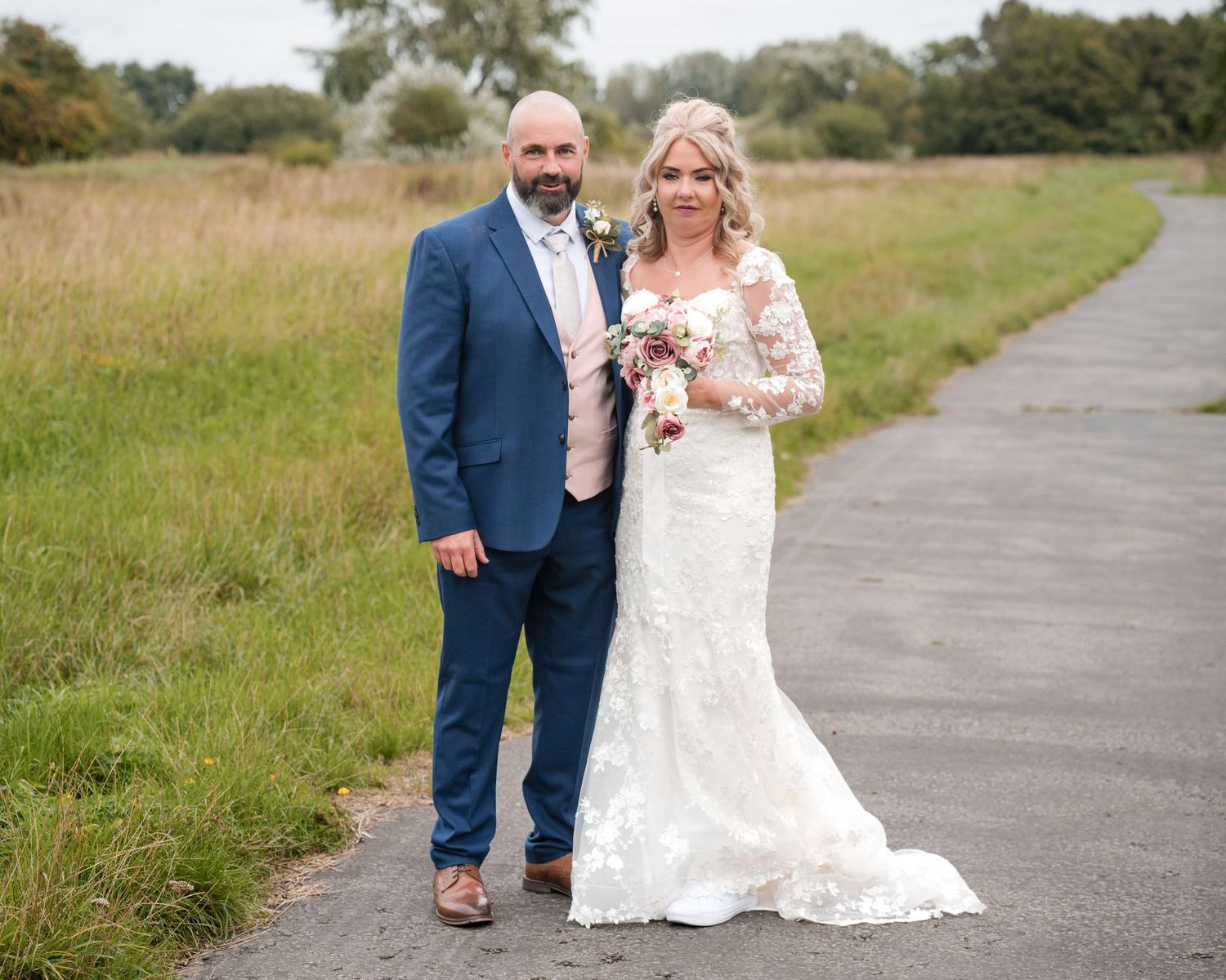 Bride in lace gown holding bouquet stands with groom in blue suit on outdoor path at countryside wedding.