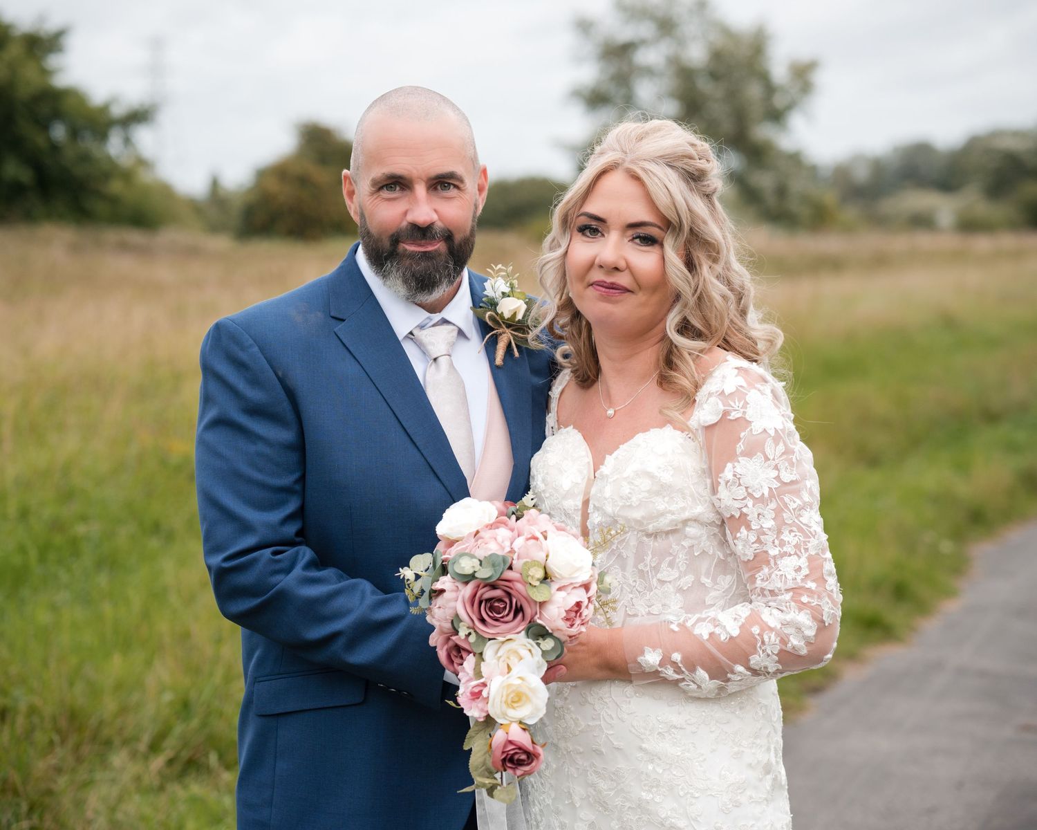 Bride and groom posing outdoors on their wedding day, she holds a pink bouquet, he wears a navy suit.