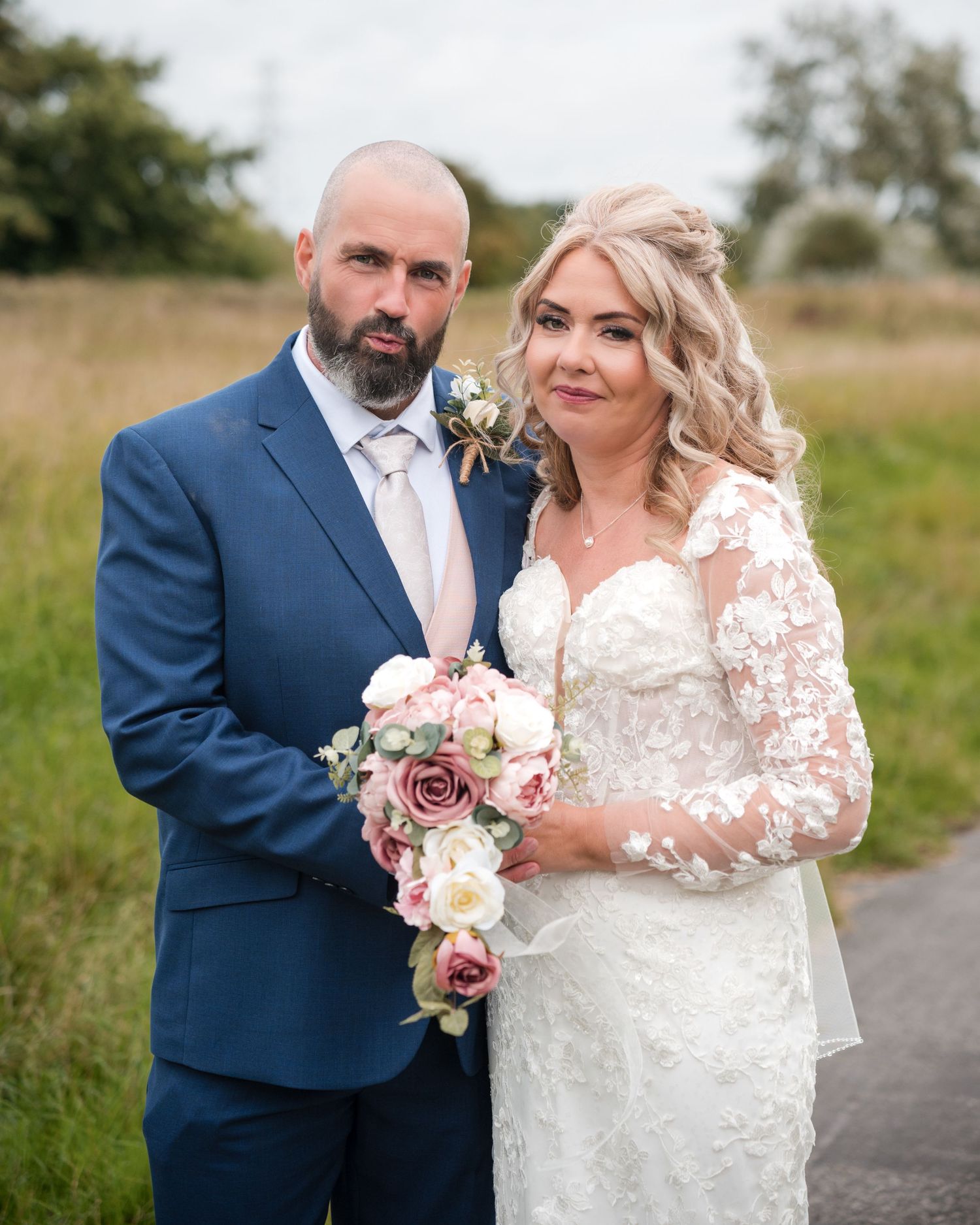 Bride and groom posing outdoors on their wedding day, she holds a pink rose bouquet wearing lace gown, he wears a navy suit.