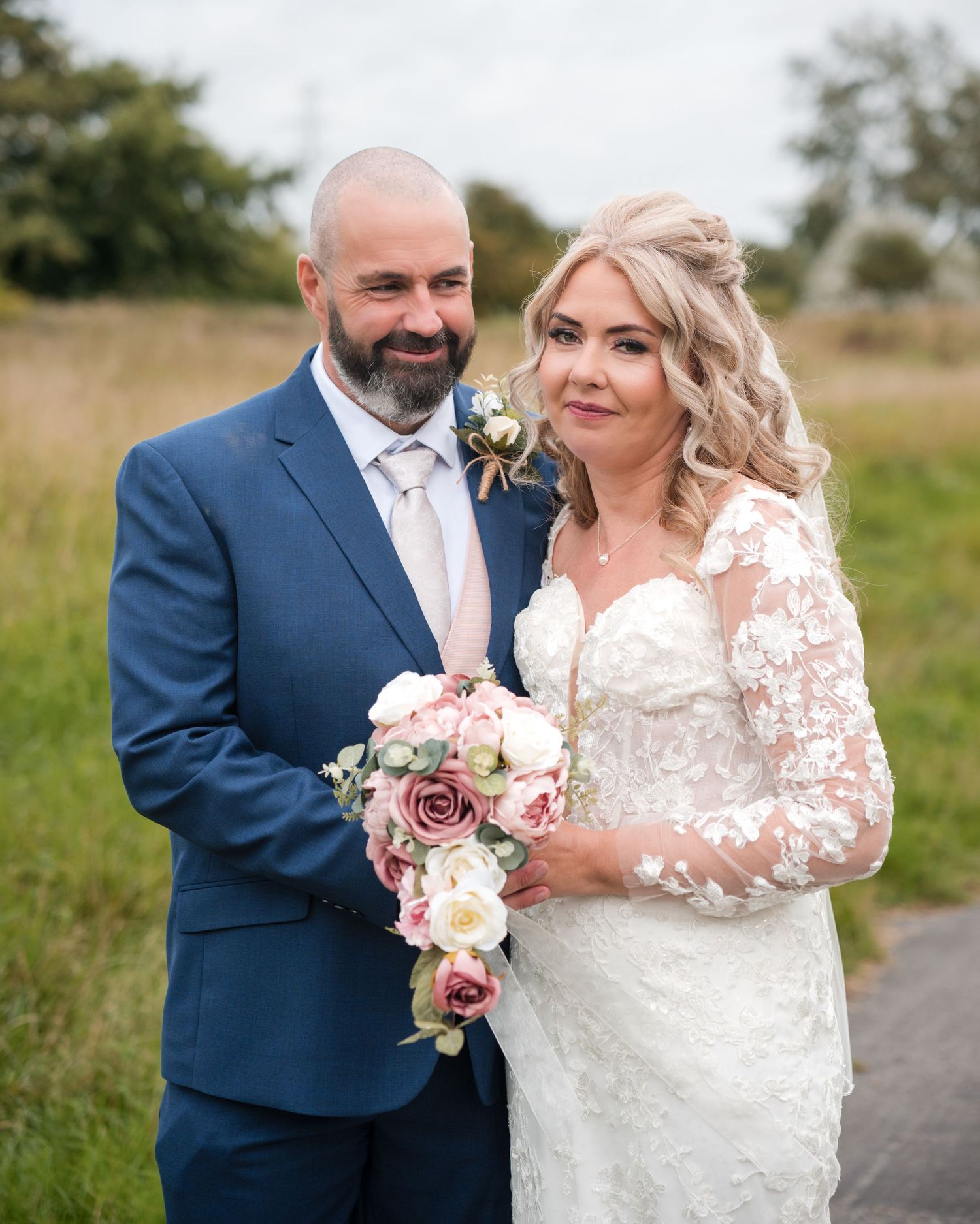 Bride in lace gown holding pink bouquet stands with groom in blue suit outdoors on their wedding day.