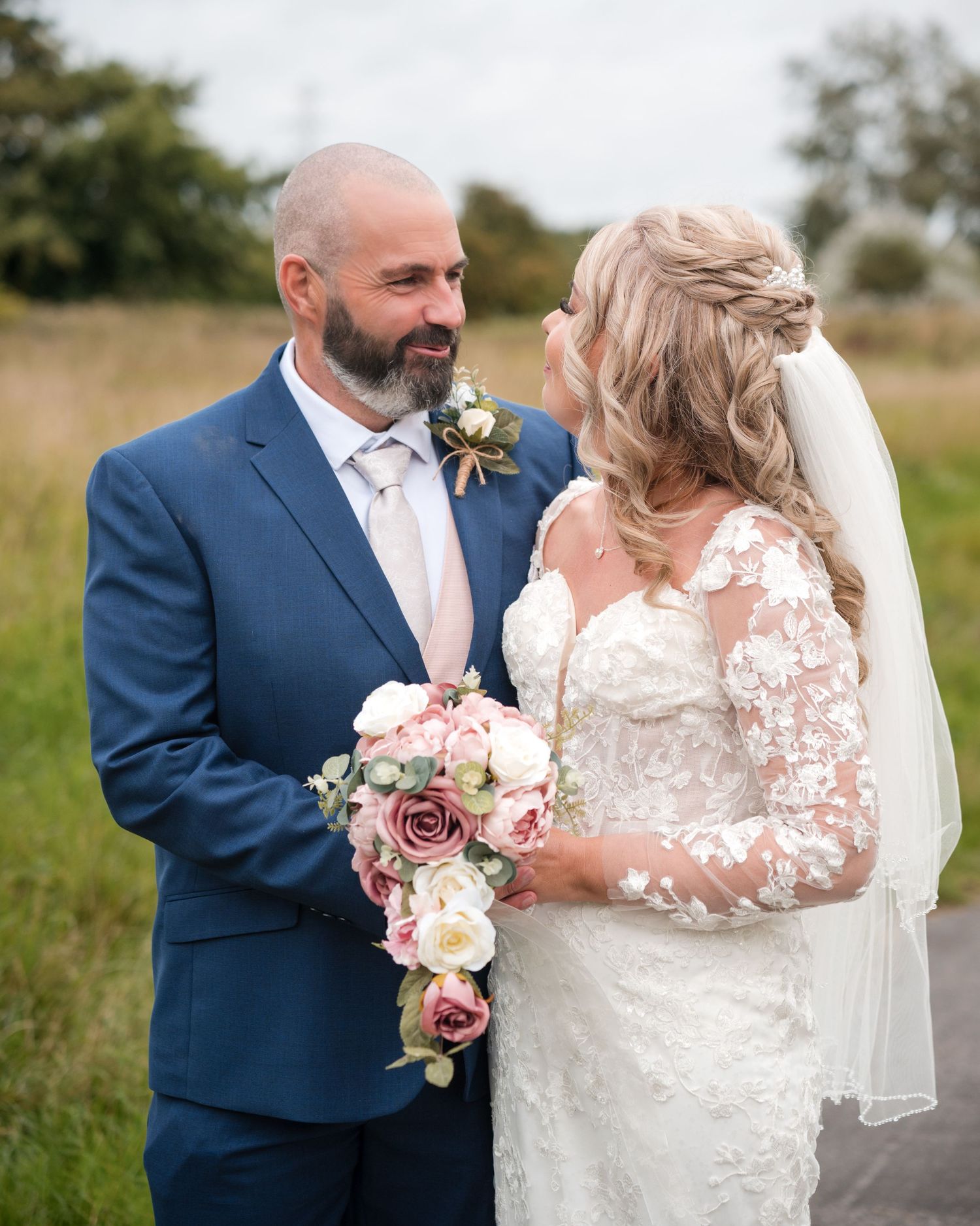 Bride and groom share a romantic moment outdoors, she holds a cascading pink and white bouquet, he wears a navy suit.