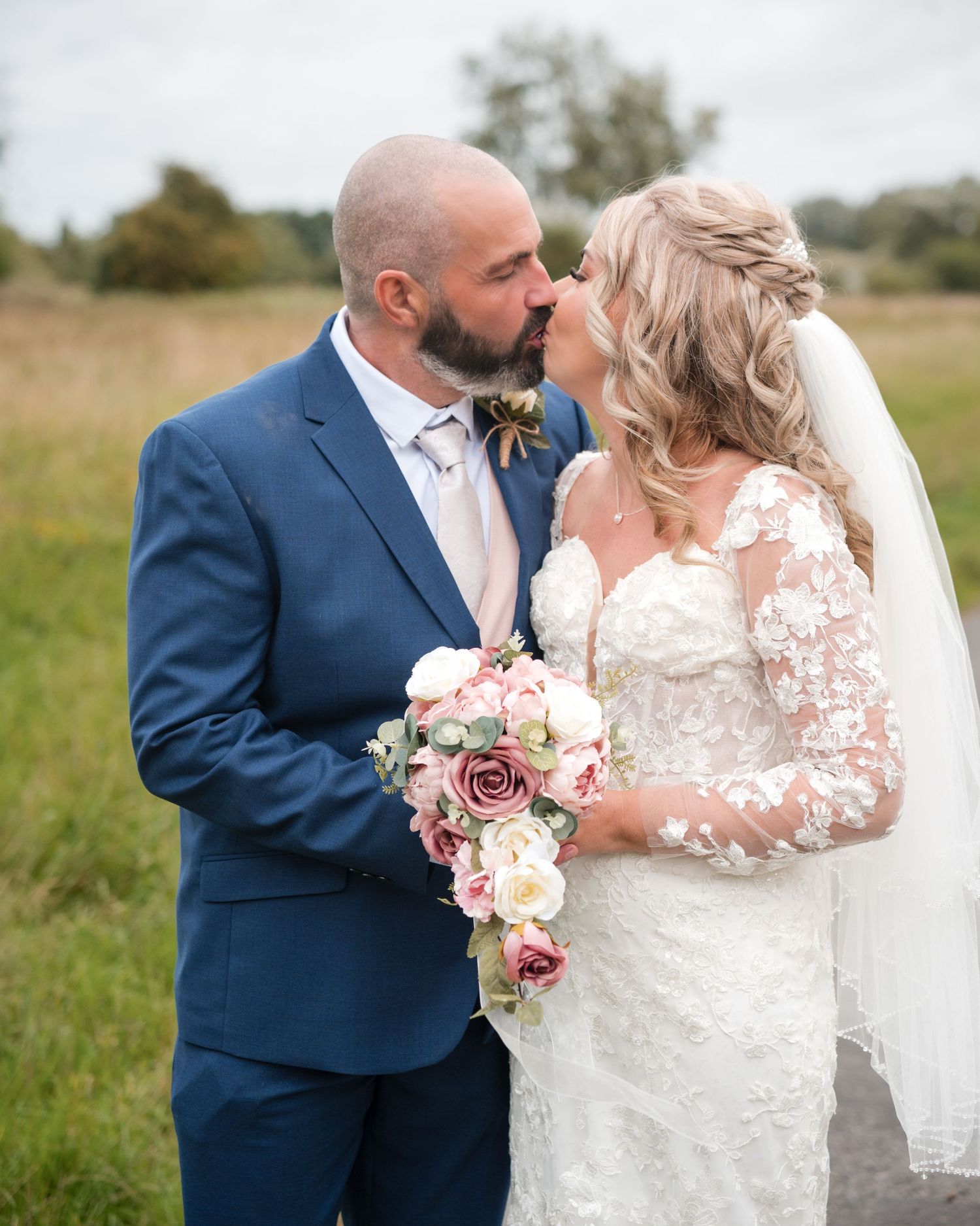A bride and groom share a romantic kiss outdoors, she holds a pink and white bouquet, wearing lace gown with veil.