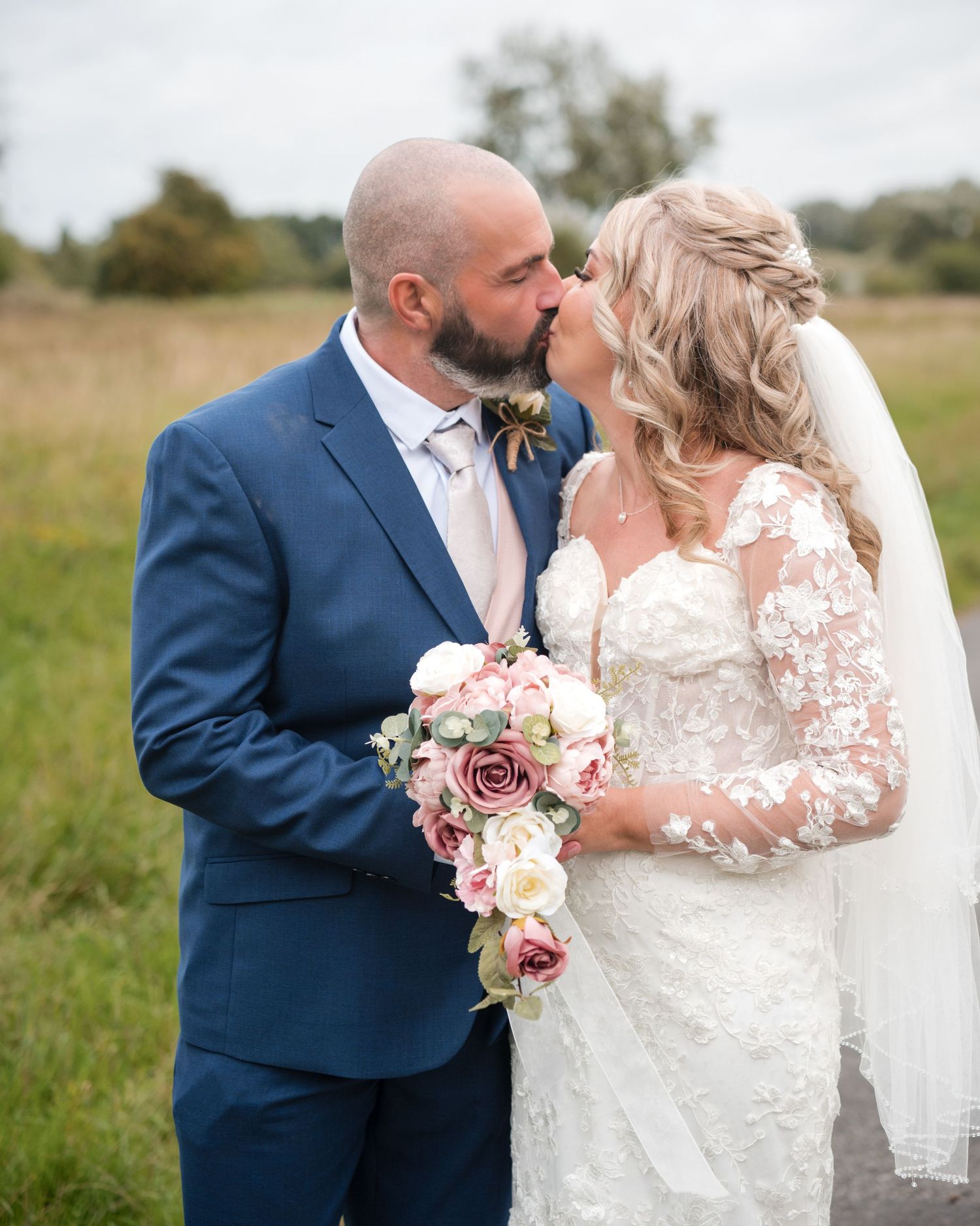 Bride and groom sharing a romantic kiss outdoors, bride holding pink and white bouquet in lace gown and veil.