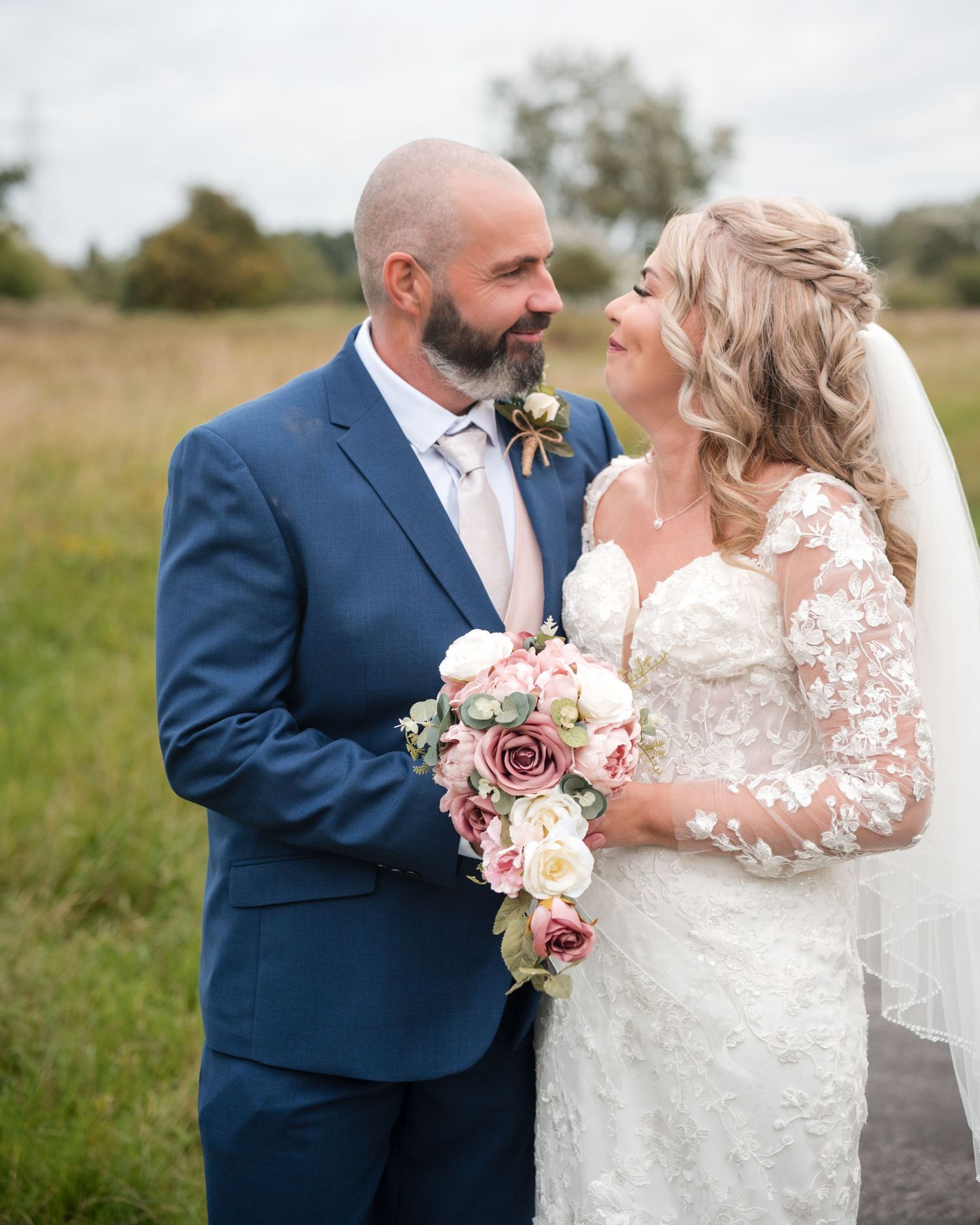 Smiling bride and groom gazing at each other outdoors, bride in lace gown holding cascading pink bouquet.