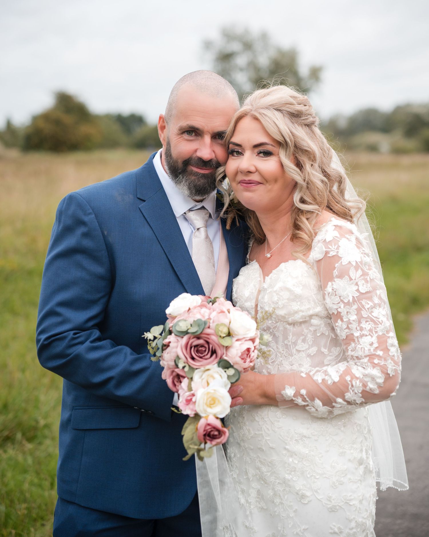 A smiling bride in lace gown holds pink bouquet beside suited groom with beard on their wedding day outdoors.