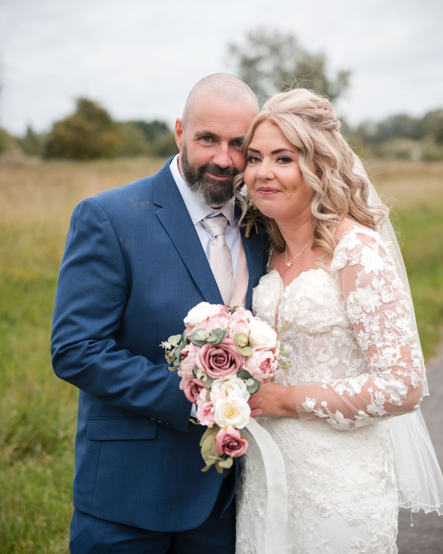 A bride and groom pose together in a field, she holds a pink bouquet, wearing lace gown, he wears a navy suit.
