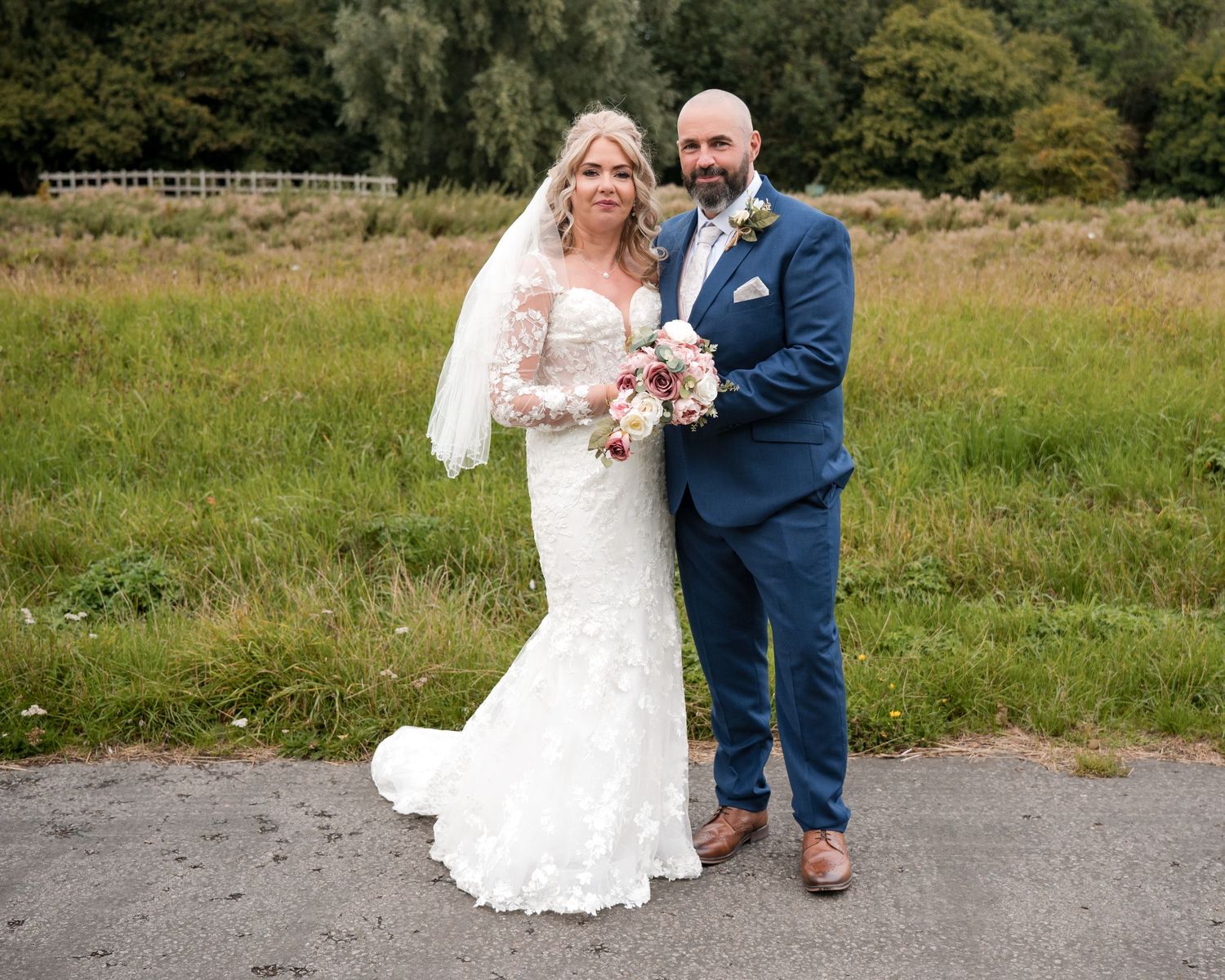 Bride in lace gown and groom in navy suit pose together outdoors on their wedding day with meadow backdrop.
