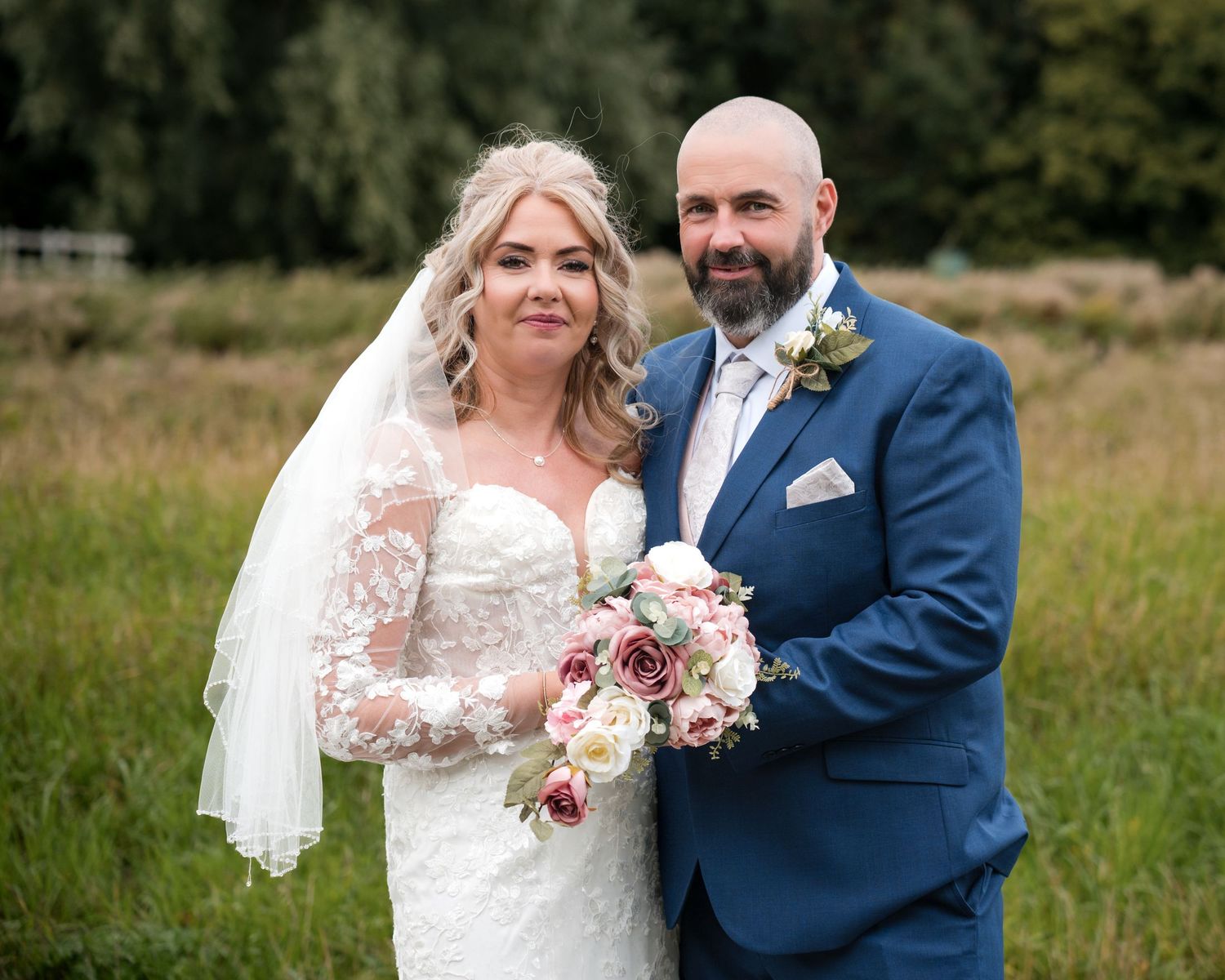 Bride and groom posing outdoors on wedding day, bride in lace gown with veil holding floral bouquet, groom in blue suit.