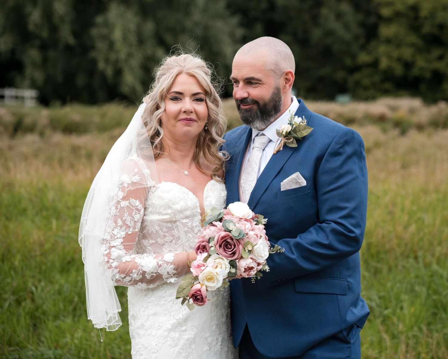 Bride in lace gown and veil holds floral bouquet beside groom in navy suit outdoors at wedding.