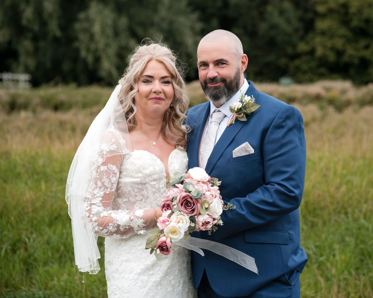 A bride in a lace gown and veil holds a floral bouquet beside a bald groom in a blue suit outdoors.