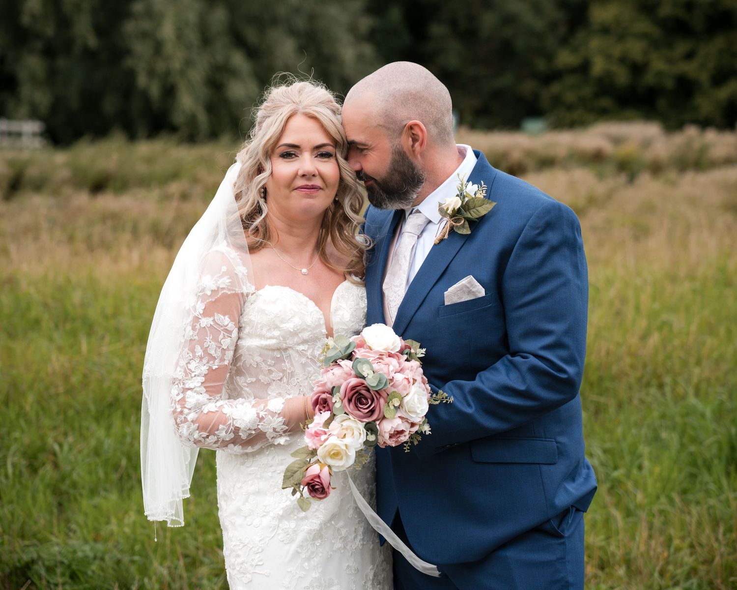Bride in lace gown with veil holds pink bouquet as groom in navy suit kisses her cheek in a field.