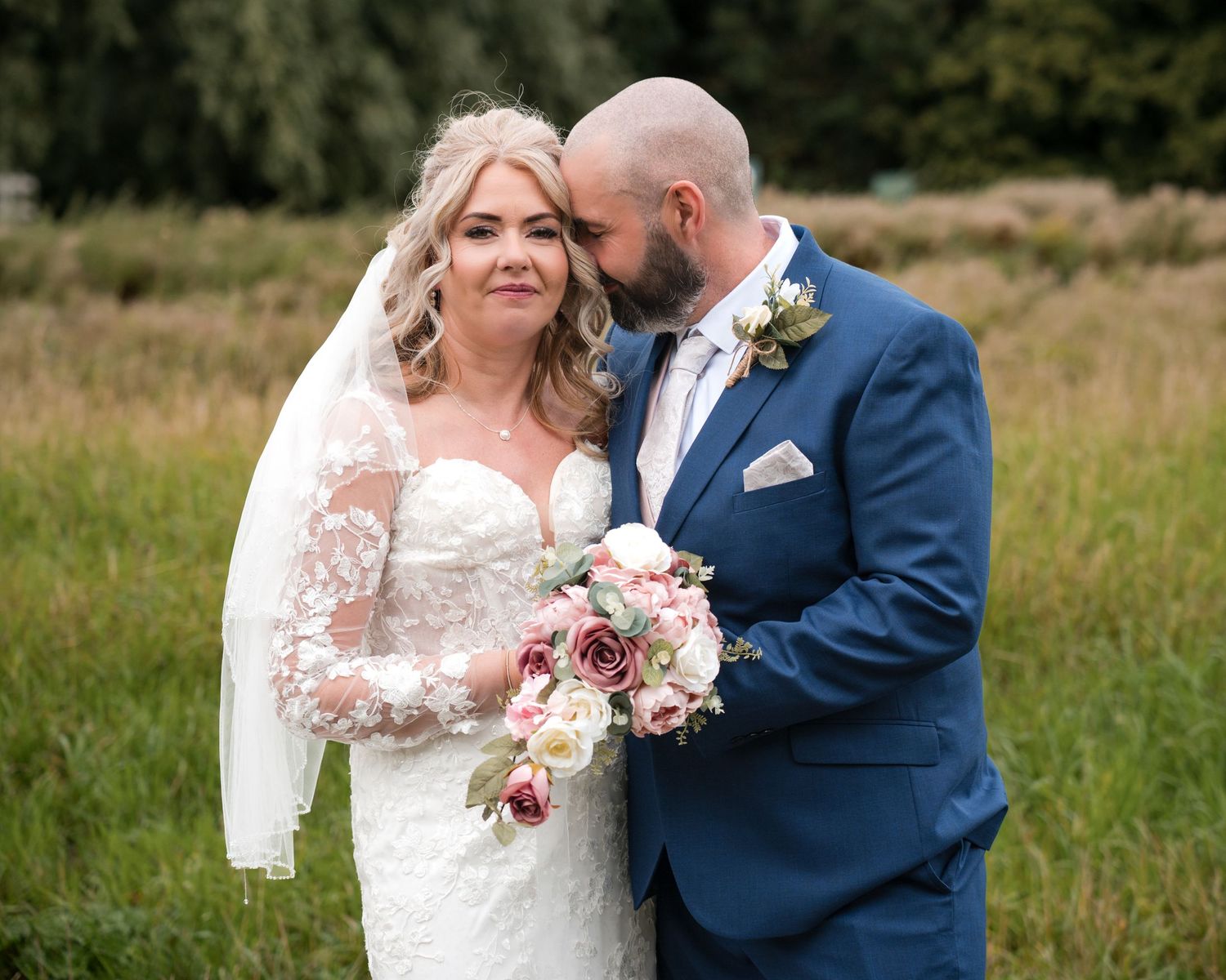 Bride in lace gown holding pink bouquet as groom in blue suit kisses her cheek outdoors.